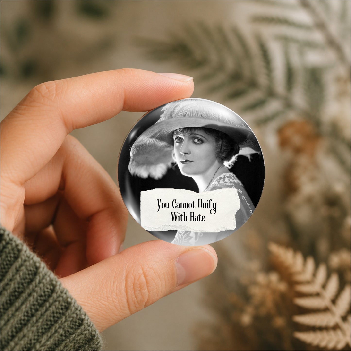 A hand holding a round pin badge featuring a black-and-white vintage portrait of a woman in a hat, with a torn-paper style overlay that reads 'You Cannot Unify With Hate.' The background is softly blurred with warm neutral tones and dried fern-like foliage.