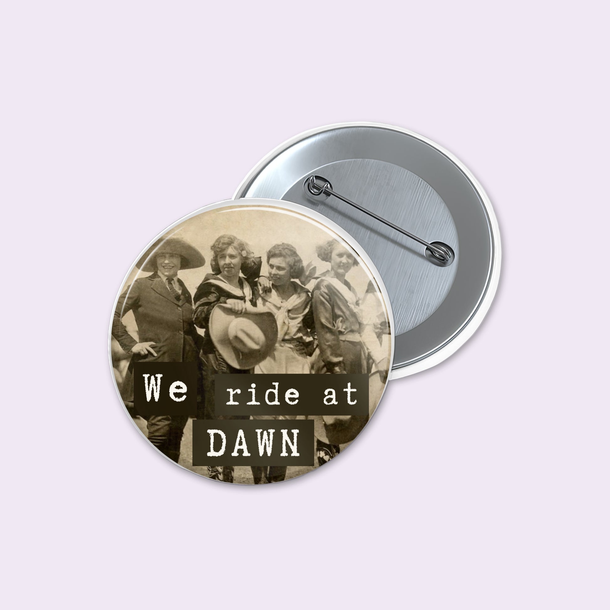 Round pinback button displays a sepia-toned vintage photograph of a group of riders with the text 'We ride at DAWN' in black.