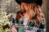 Woman wearing an upcycled book page necklace, green plaid flannel, and a black hat holding a mug outdoors with a blurred background