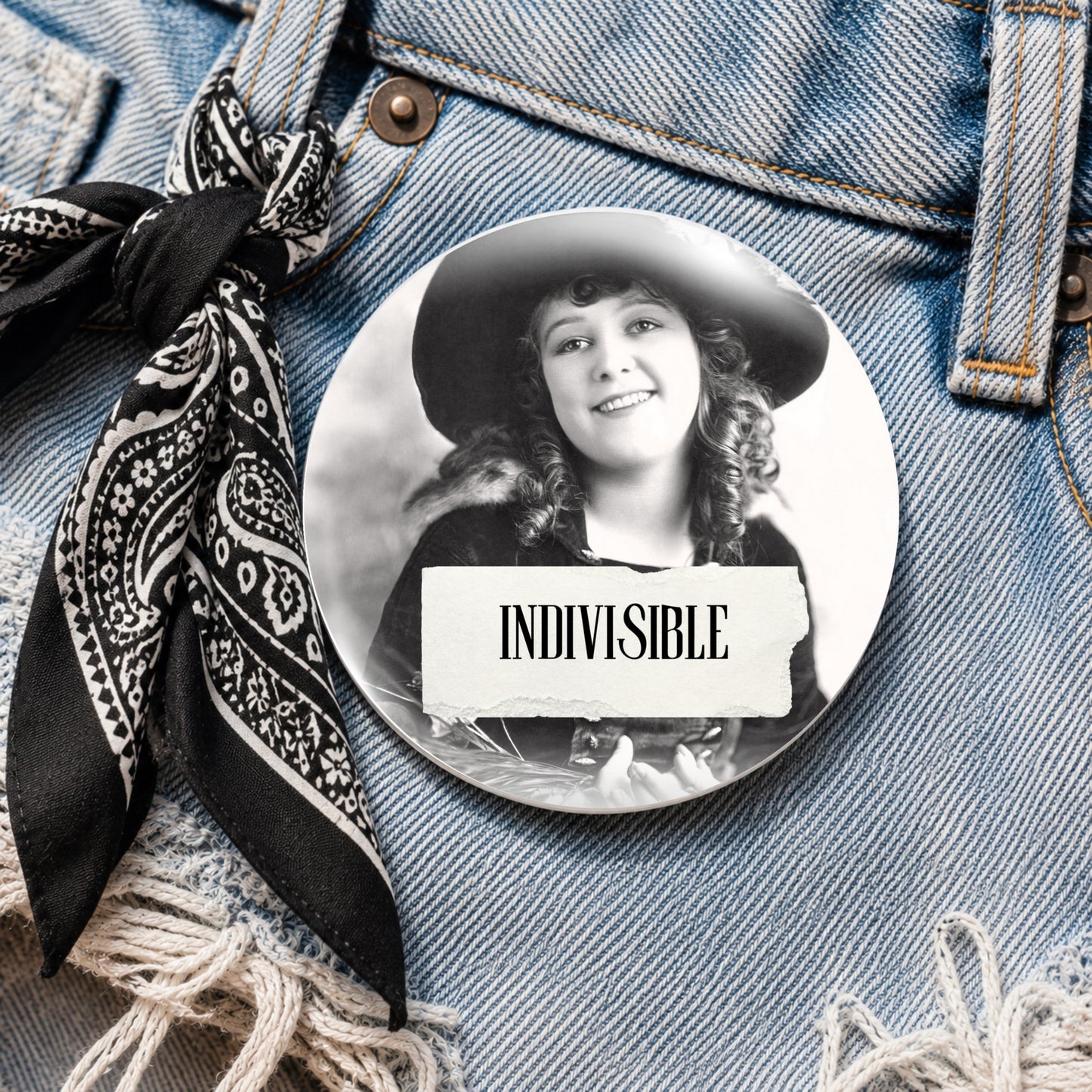 A round pinback button featuring a black and white portrait of a woman in a hat with a torn paper label reading 'INDIVISIBLE', displayed on blue denim jeans next to a black paisley bandana.