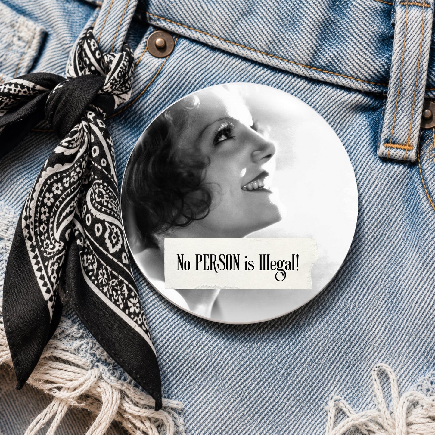 A round pin badge featuring a black and white portrait of a woman with the text 'No PERSON is Illegal!' placed on a blue denim jacket next to a black and white paisley neck scarf.