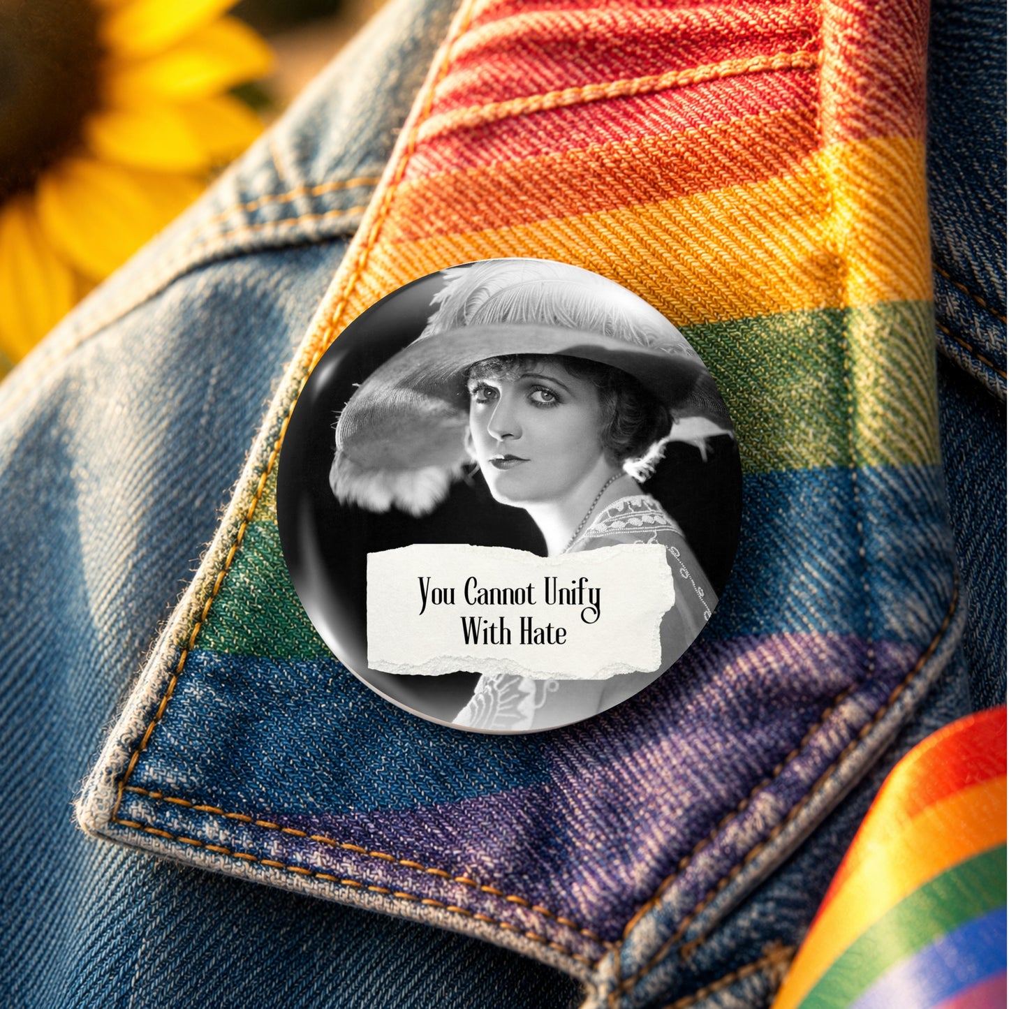A round pin-back button badge featuring a black-and-white portrait of a woman in a hat with the text 'You Cannot Unify With Hate,' placed on a denim jacket with a rainbow-striped fabric accent.