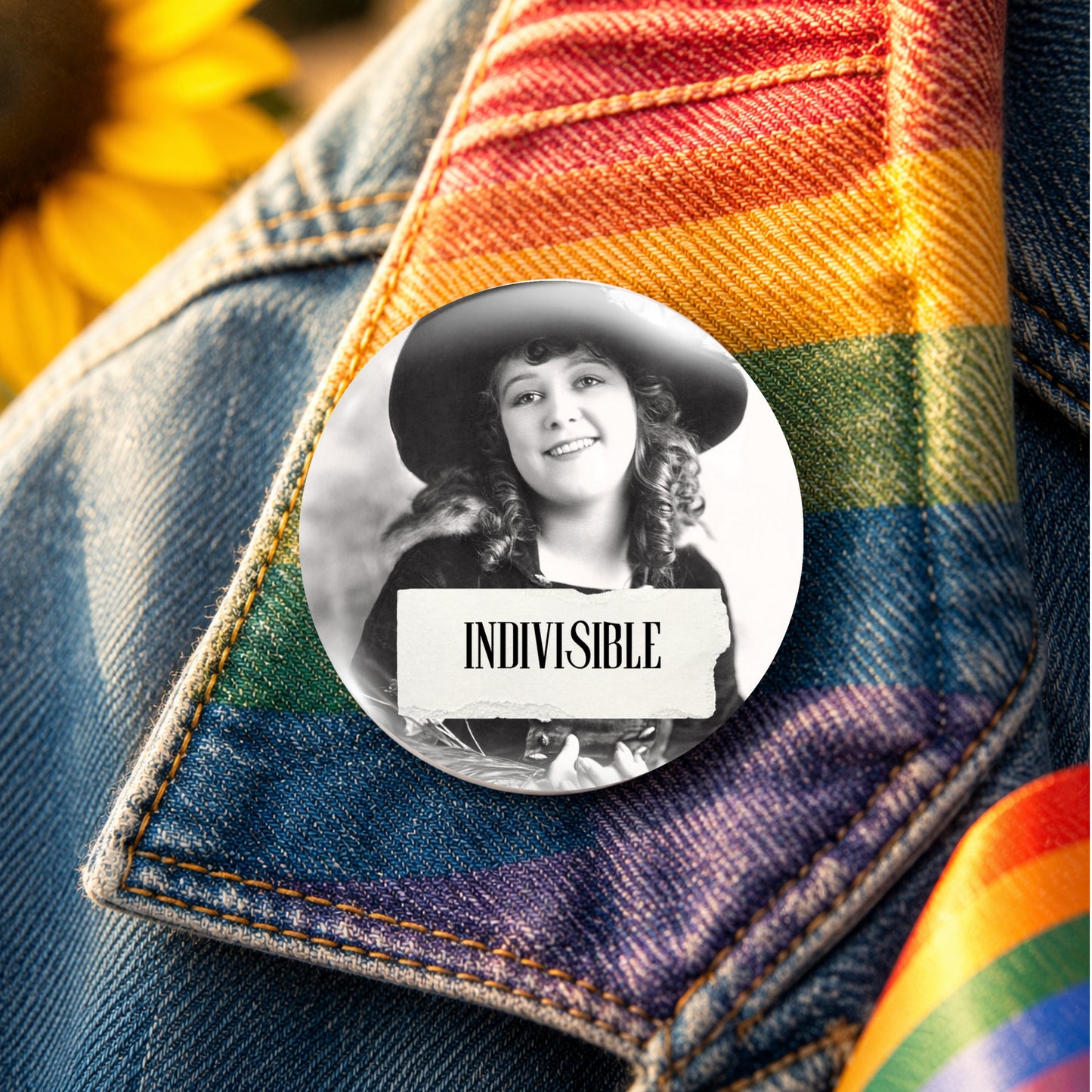 A round pin badge featuring a black-and-white photo of a smiling woman in a hat with the word 'INDIVISIBLE' on a torn-paper style label, pinned onto a denim jacket over a rainbow-striped fabric.