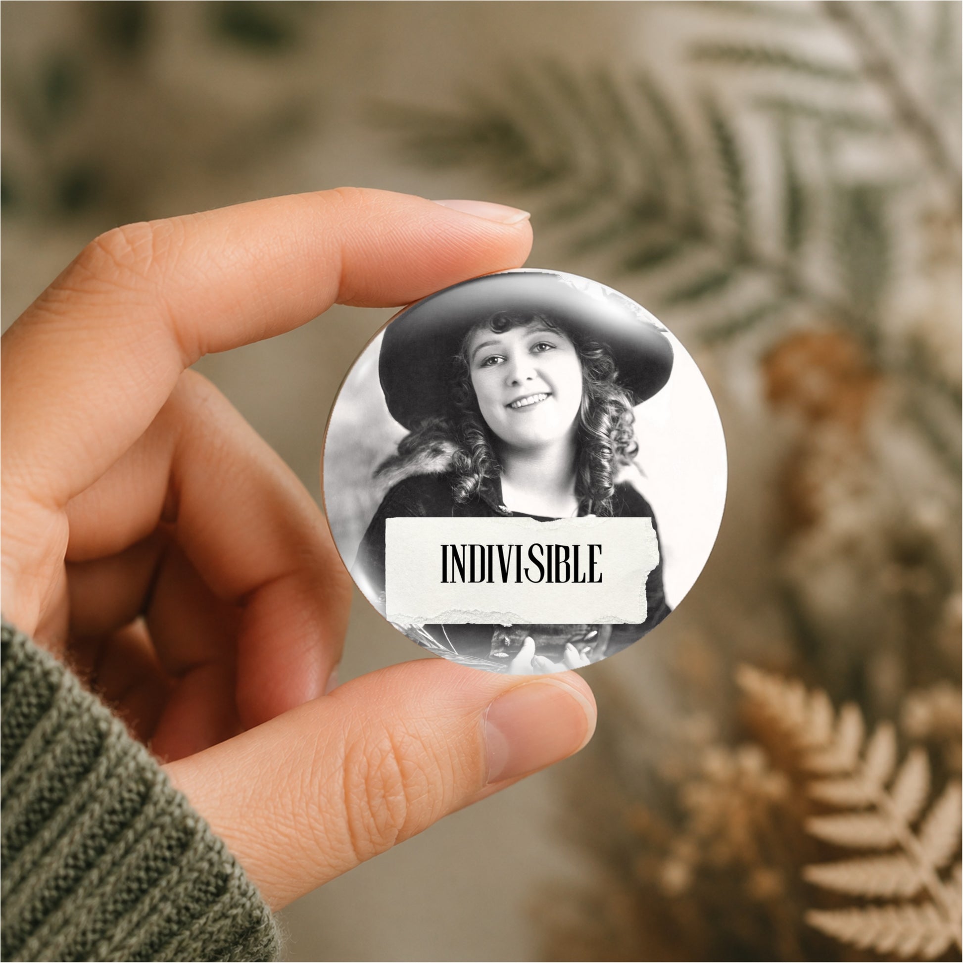 A hand holds a round pin-back button featuring a black-and-white portrait of a woman in a hat with a torn-paper style banner reading 'INDIVISIBLE'. The background is softly blurred with dried fern-like foliage.