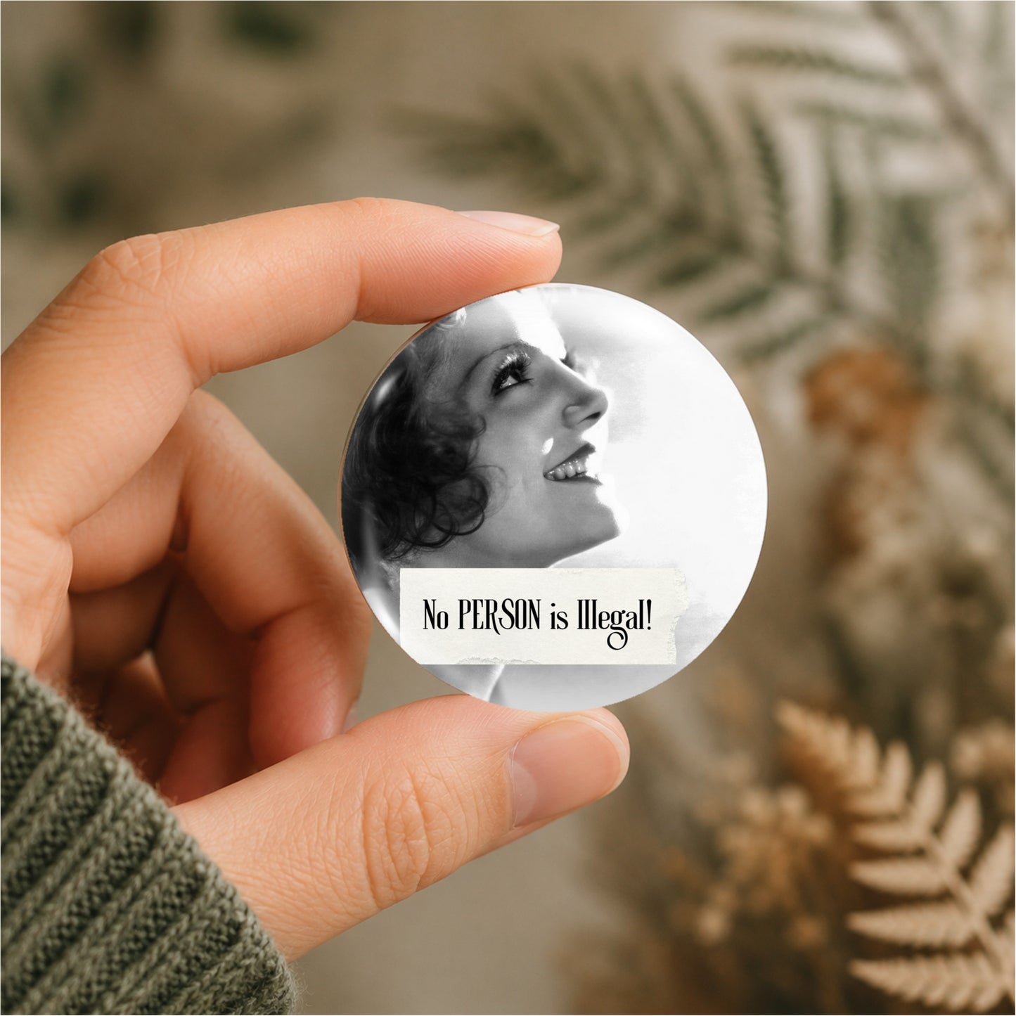 A hand holding a round pin badge featuring a black and white portrait of a smiling woman and the text 'No PERSON is Illegal!'. The background is a soft-focus beige and brown dried fern setting.