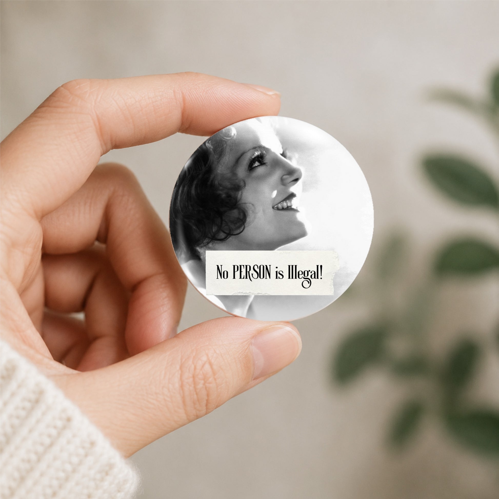 A hand wearing a white knit sweater holds a round pin badge featuring a black and white photo of a smiling woman and the text 'No PERSON is illegal!'. The background is a soft, out-of-focus beige wall with green leaves.