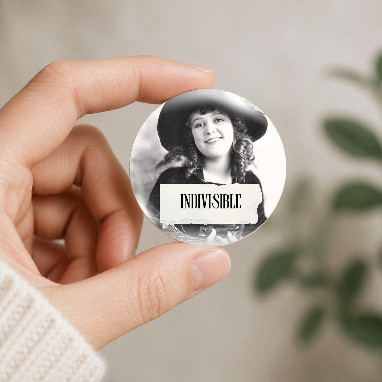 A hand holds a round pin-back button featuring a black-and-white portrait of a smiling woman in a hat with the word 'INDIVISIBLE' on a white banner, against a softly blurred indoor background.