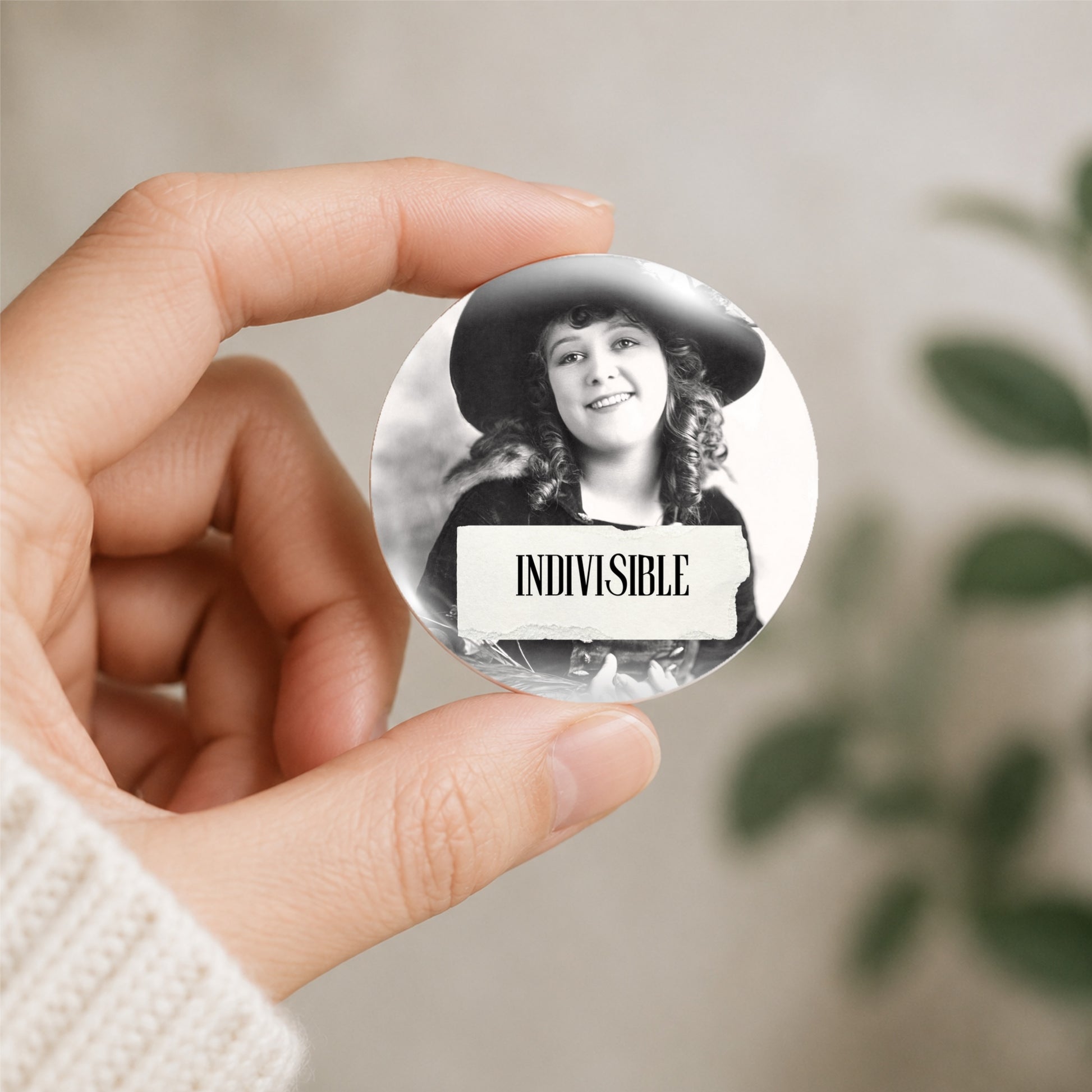 A hand holds a round pin-back button featuring a black-and-white portrait of a smiling woman in a hat with the word 'INDIVISIBLE' on a white banner, against a softly blurred indoor background.