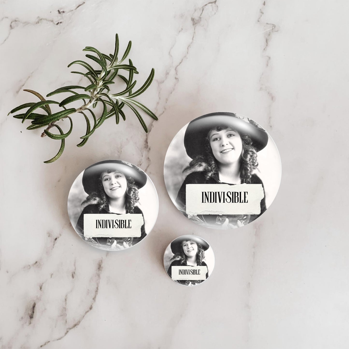 Three round pin-back buttons featuring a black-and-white portrait of a woman in a hat with a torn-paper label reading 'INDIVISIBLE,' arranged on a white marble surface beside a green rosemary sprig.
