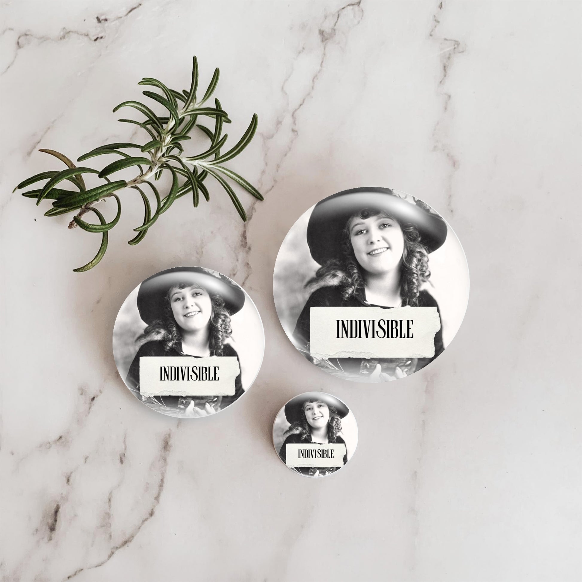 Three round pin-back buttons featuring a black-and-white portrait of a woman in a hat with a torn-paper label reading 'INDIVISIBLE,' arranged on a white marble surface beside a green rosemary sprig.
