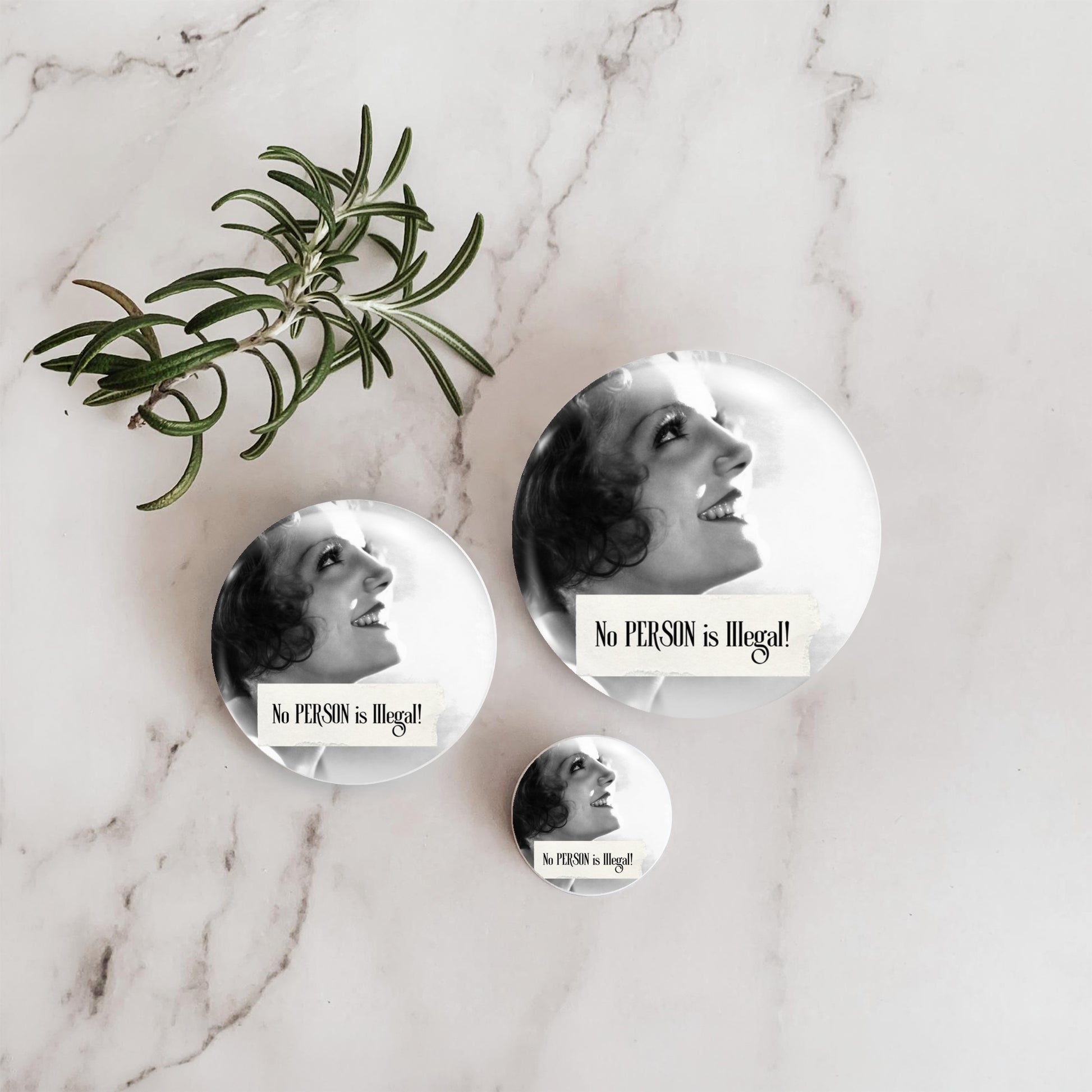 Three round pin-back buttons with a black-and-white portrait of a smiling woman and the text 'No PERSON is illegal!' arranged on a white marble surface beside a green rosemary sprig.