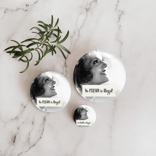 Three round pin-back buttons with a black-and-white portrait of a smiling woman and the text 'No PERSON is illegal!' arranged on a white marble surface beside a green rosemary sprig.