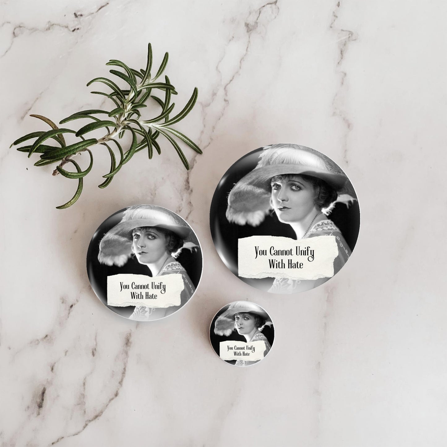 Three round pin-back buttons featuring a black-and-white vintage portrait of a woman in a hat, arranged on a white marble surface with a green herb sprig. The buttons read 'You Cannot Unify With Hate' and 'I Cannot Unify With Hate.