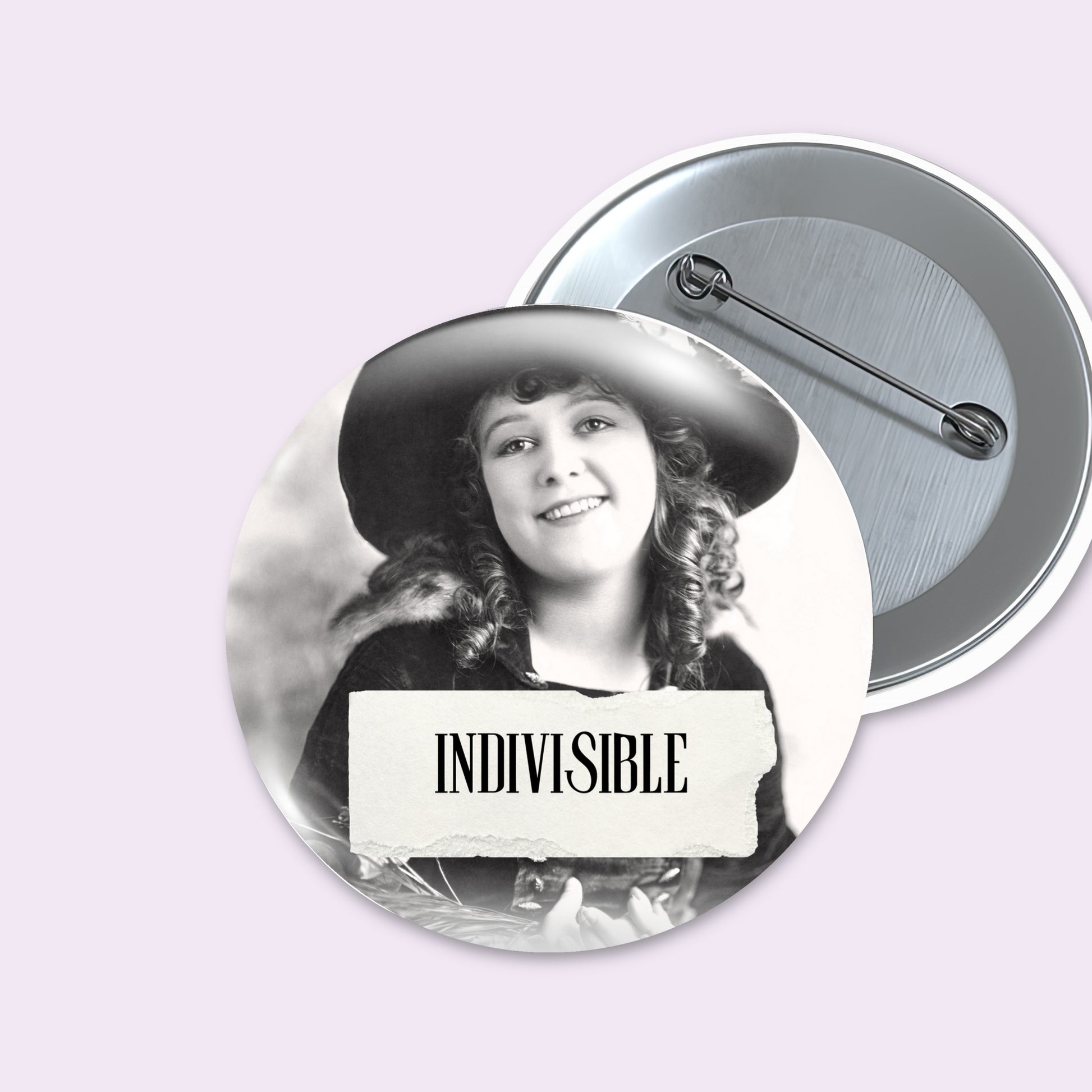 Two round pinback buttons on a pale lavender background. The front button shows a black-and-white portrait of a smiling woman in a hat with a torn-paper style banner reading 'INDIVISIBLE.