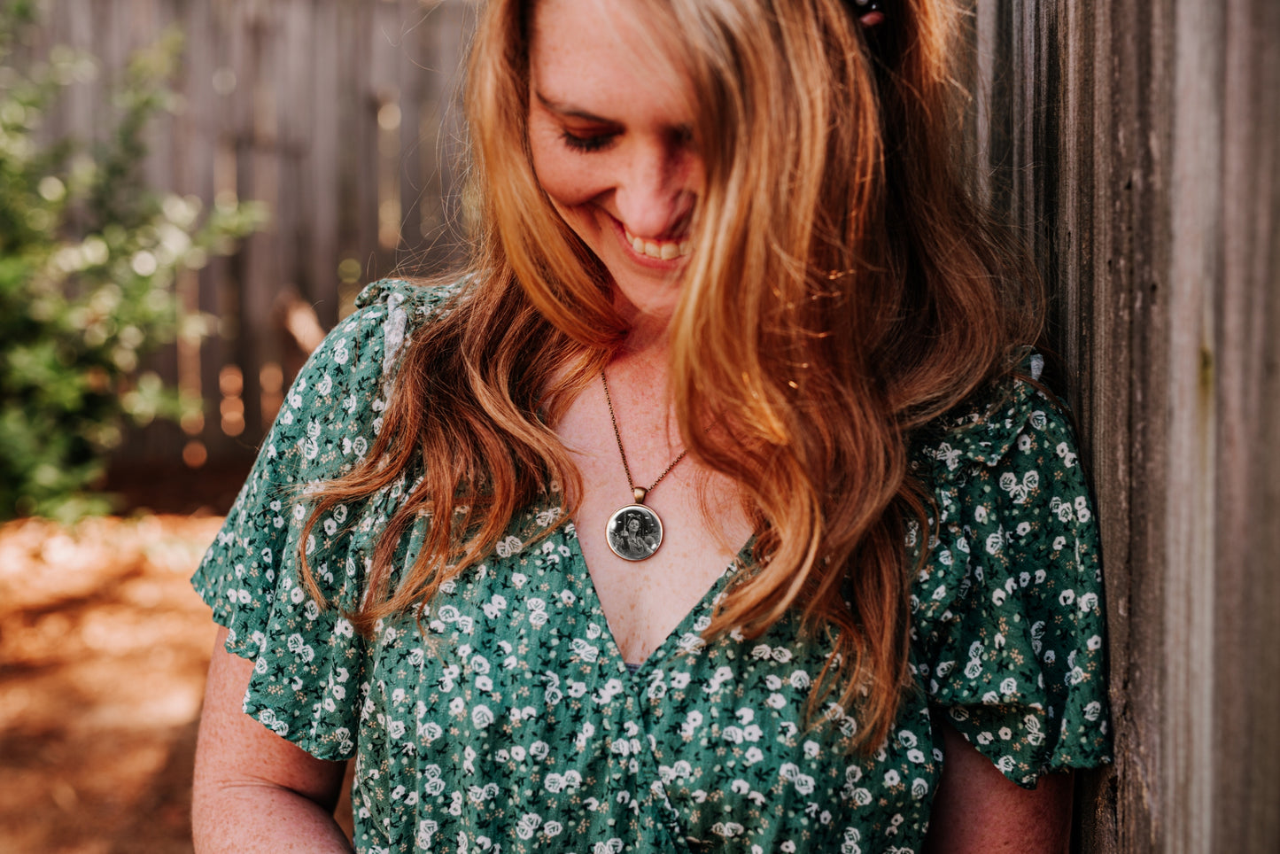 A woman with long wavy light brown hair leans against a weathered wooden fence, wearing a green short-sleeve V-neck dress with a white floral pattern and a silver pendant necklace.