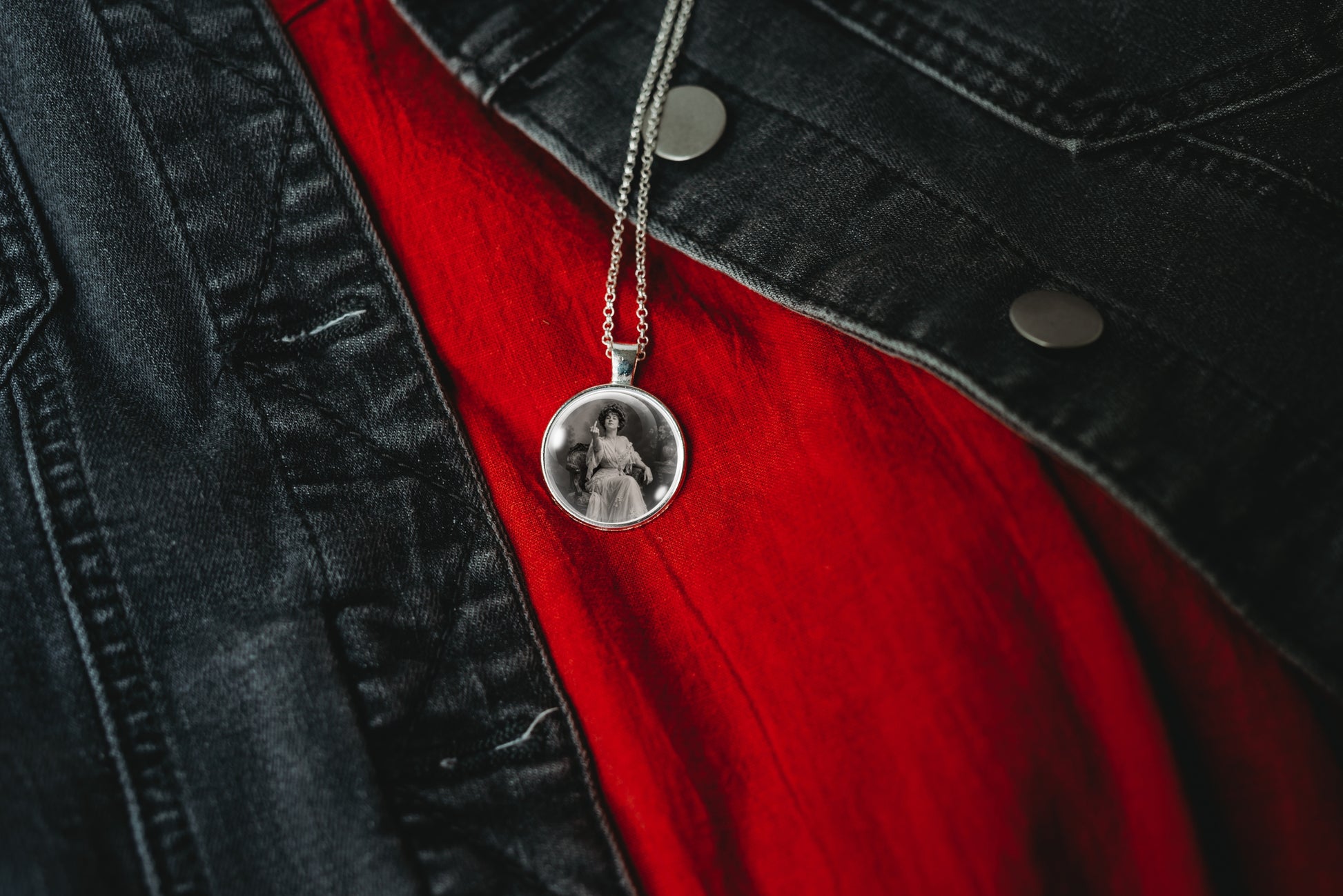 A silver-plated pendant necklace with a custom circular photo insert hanging from a chain, styled over a red shirt and black denim jacket.