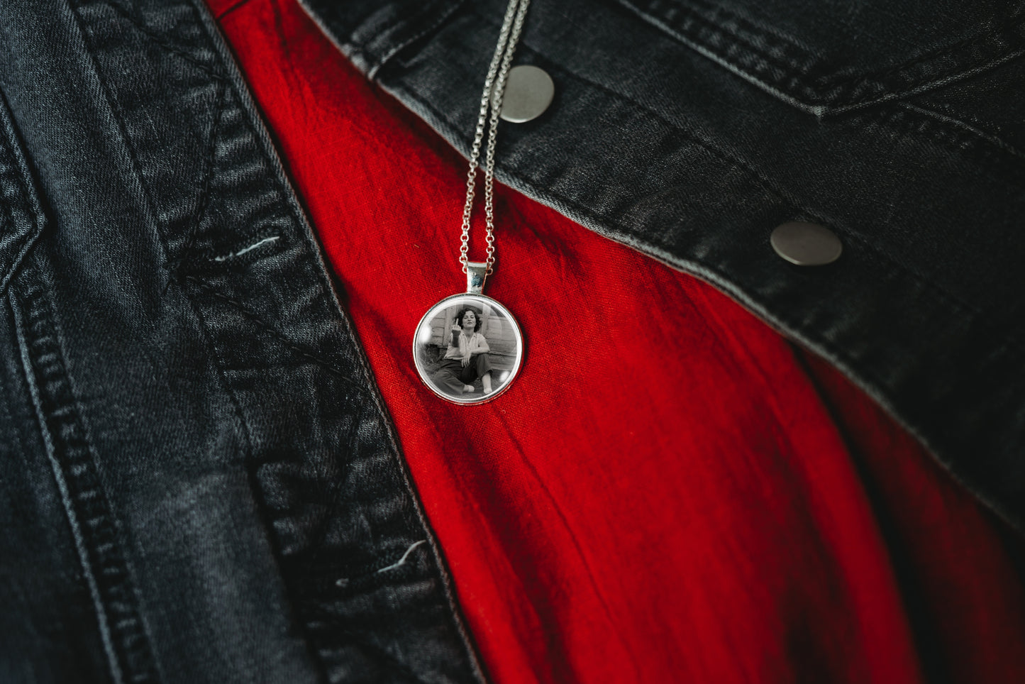 A silver round pendant necklace with a black-and-white photo printed inside, hanging from a silver chain over a red shirt and black denim jacket.