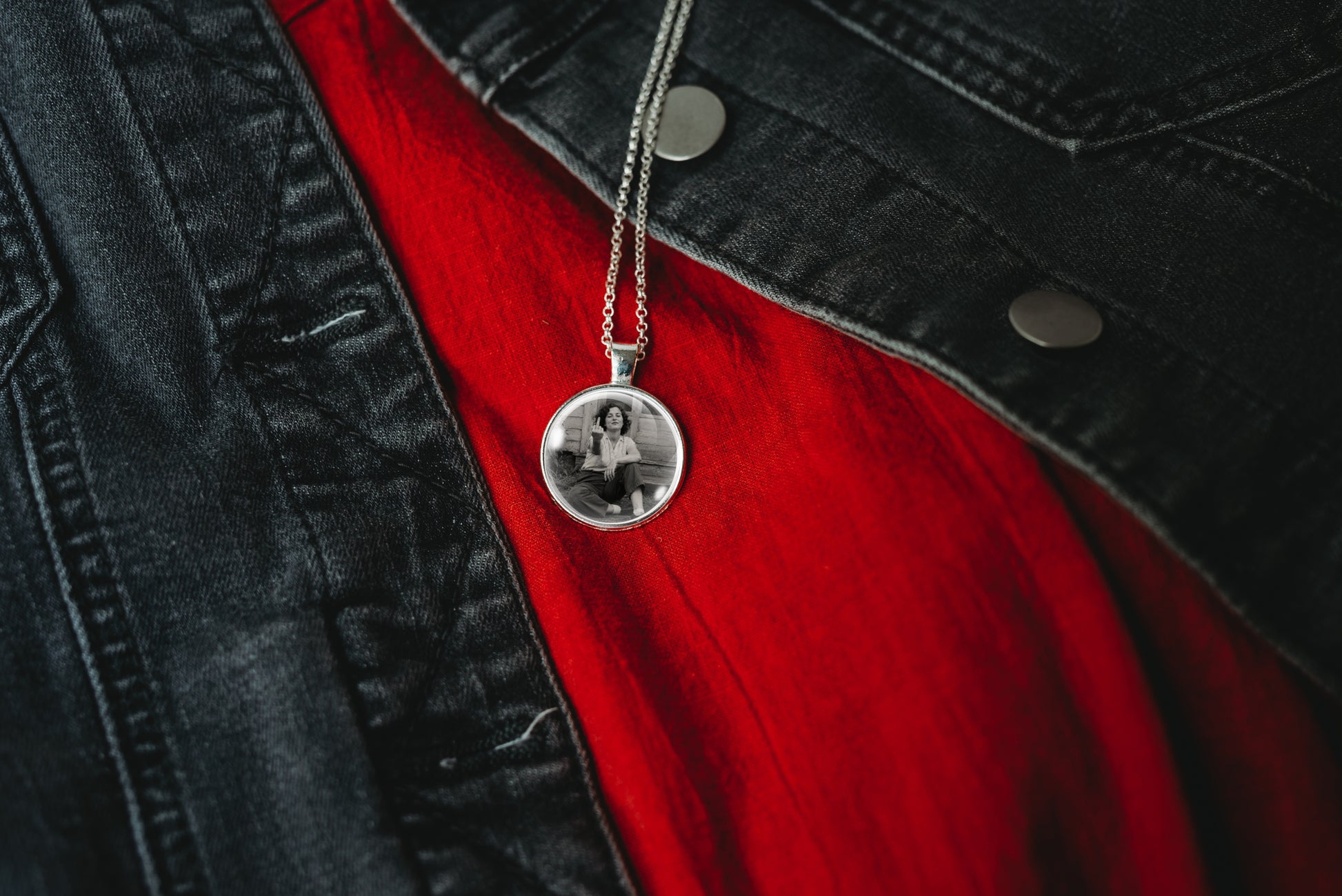 A silver round pendant necklace with a black-and-white photo printed inside, hanging from a silver chain over a red shirt and black denim jacket.