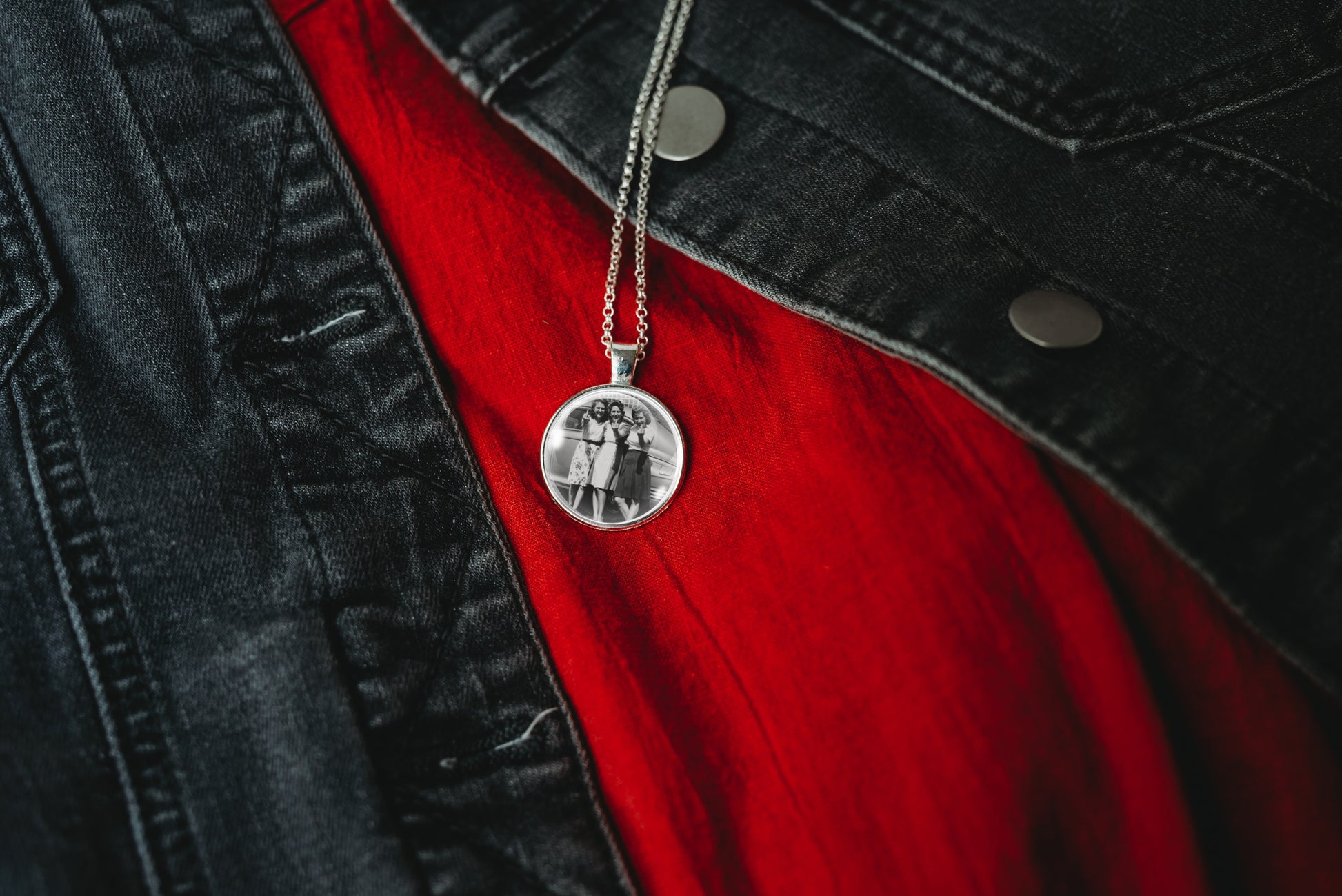 A silver round pendant necklace with a black-and-white photo of three women, displayed on a red shirt under a black denim jacket.