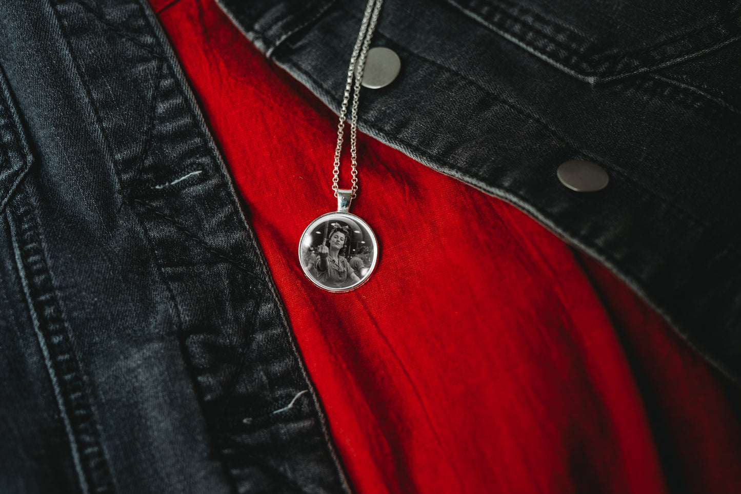 A silver pendant necklace with a circular black-and-white portrait medallion, hanging from a chain over a bright red shirt and dark denim jacket.