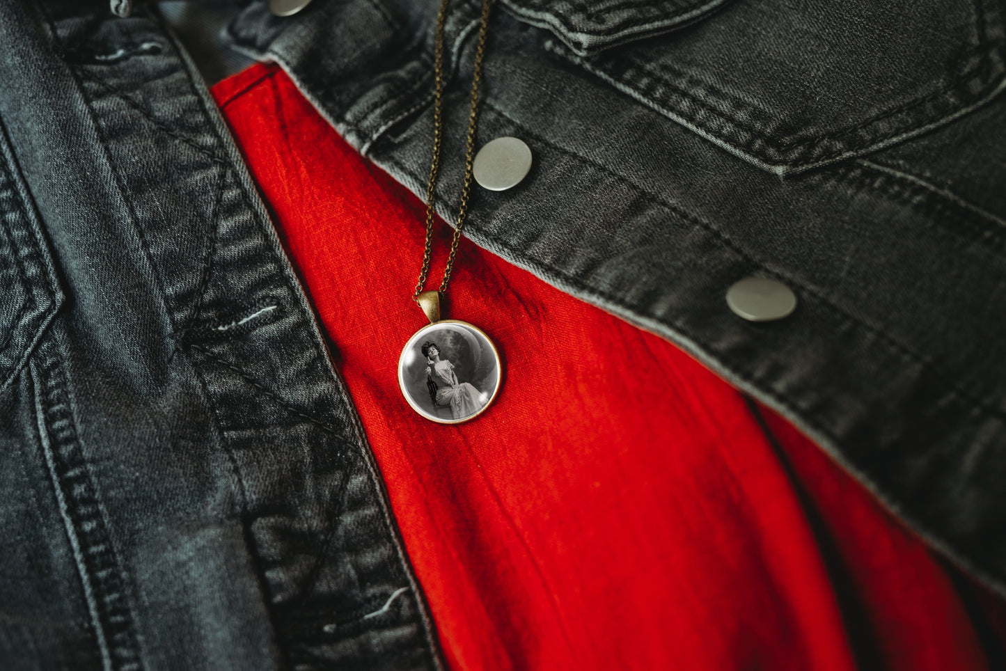 A close-up of a circular pendant necklace on a dark chain, resting on a bright red shirt and a black denim jacket.