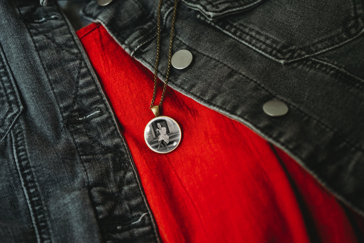 A close-up of a circular pendant necklace featuring a black-and-white portrait image, hanging from a dark chain over a bright red shirt and a dark gray denim jacket.
