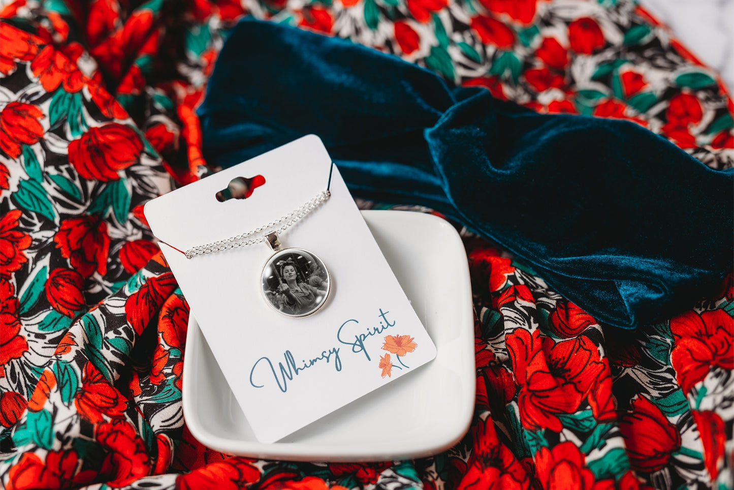 A photo necklace on a delicate chain, displayed on a white card labeled 'Whimsy Spirit' resting on a small white dish. The necklace is placed on a dark navy velvet bow over a red and green floral patterned fabric background.