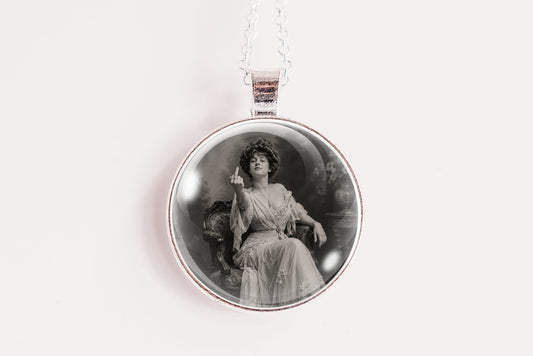 A silver-toned circular pendant necklace featuring a black and white vintage portrait of a seated woman, holding up her middle finger, suspended from a thin silver chain against a plain white background.