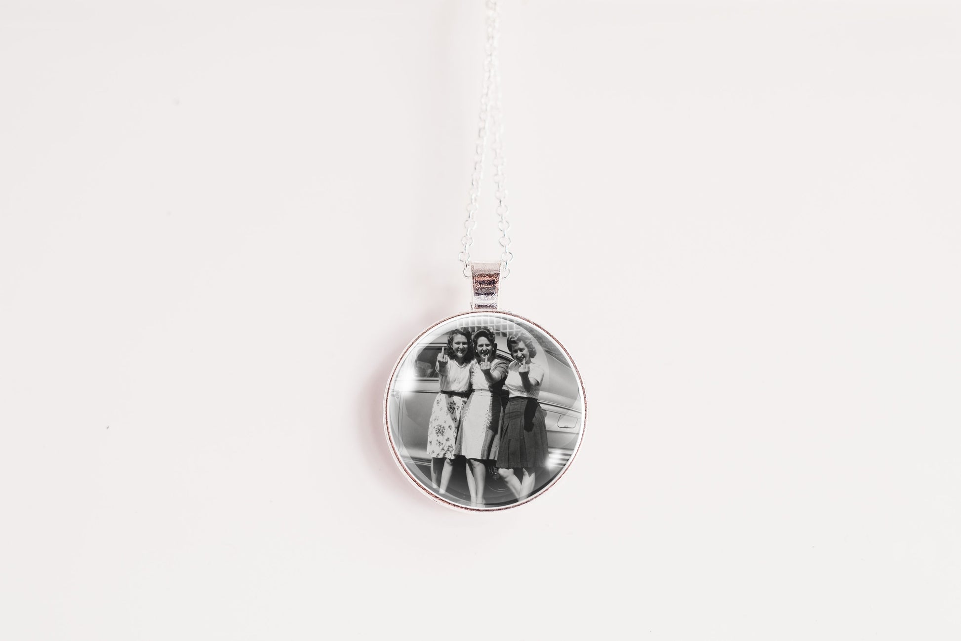 A silver round pendant necklace featuring a black and white photograph of three women, suspended from a white cord against a plain white background.