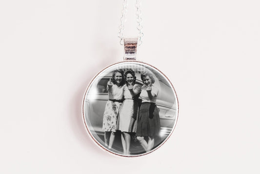 A silver round pendant necklace featuring a black and white photograph of three women, suspended from a white cord against a plain white background.