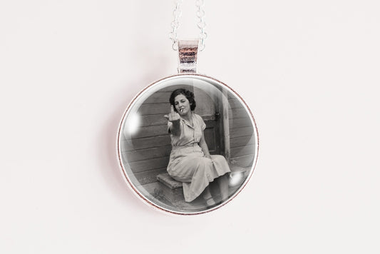 A silver circular pendant necklace featuring a black and white vintage photograph of a woman sitting on steps, suspended from a silver chain against a plain white background.