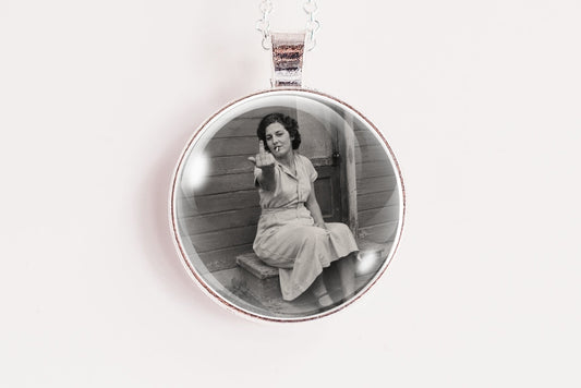 A silver circular pendant necklace featuring a black and white vintage photograph of a woman sitting on steps, suspended from a white cord against a plain white background.