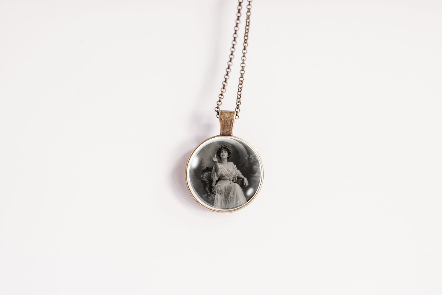 A bronze-plated circular pendant necklace featuring a black and white vintage portrait of a seated woman, holding up her middle finger, suspended from a thin silver chain against a plain white background.