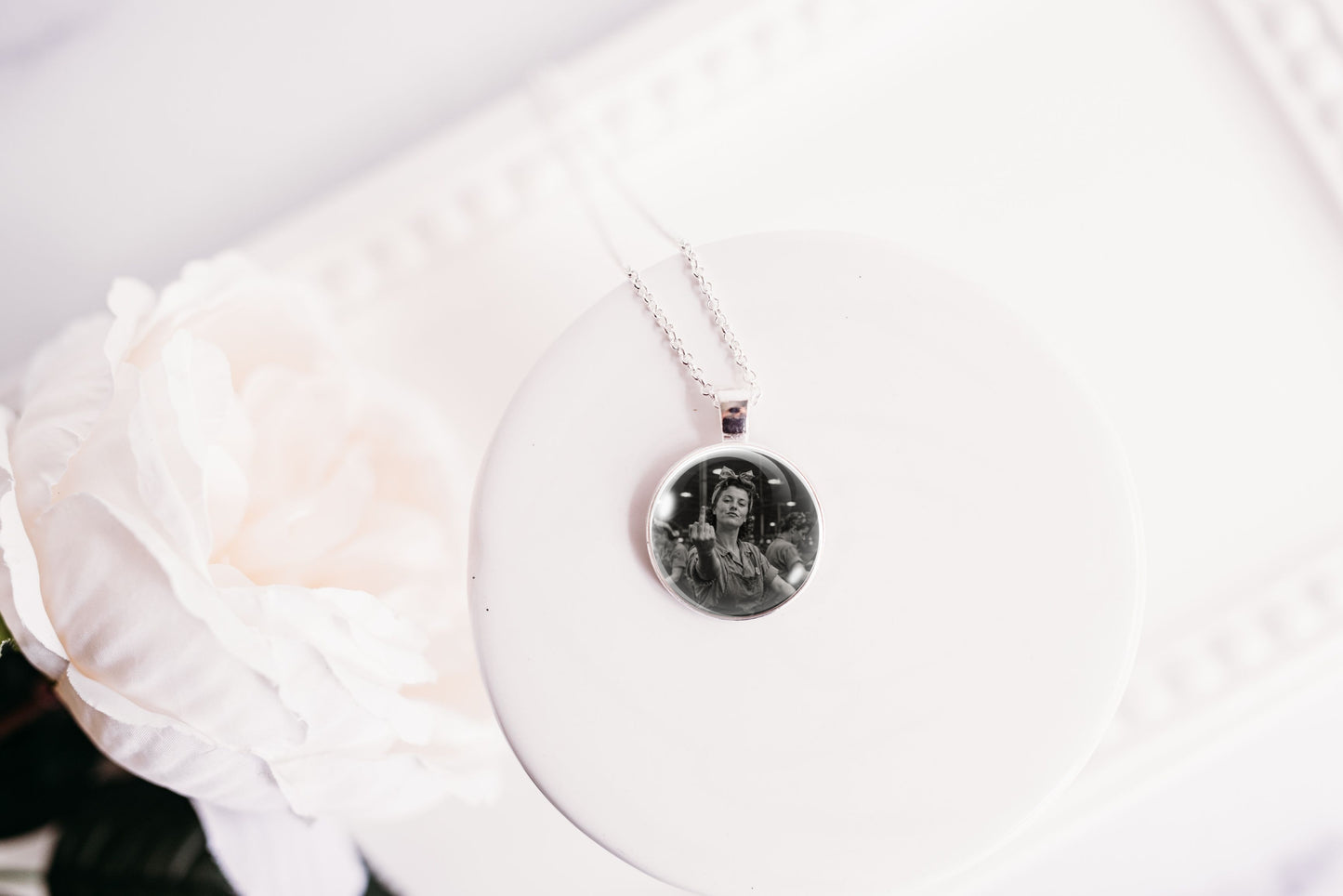A silver necklace with a round pendant displaying a black and white portrait photo, resting on a white circular display stand next to a white rose.