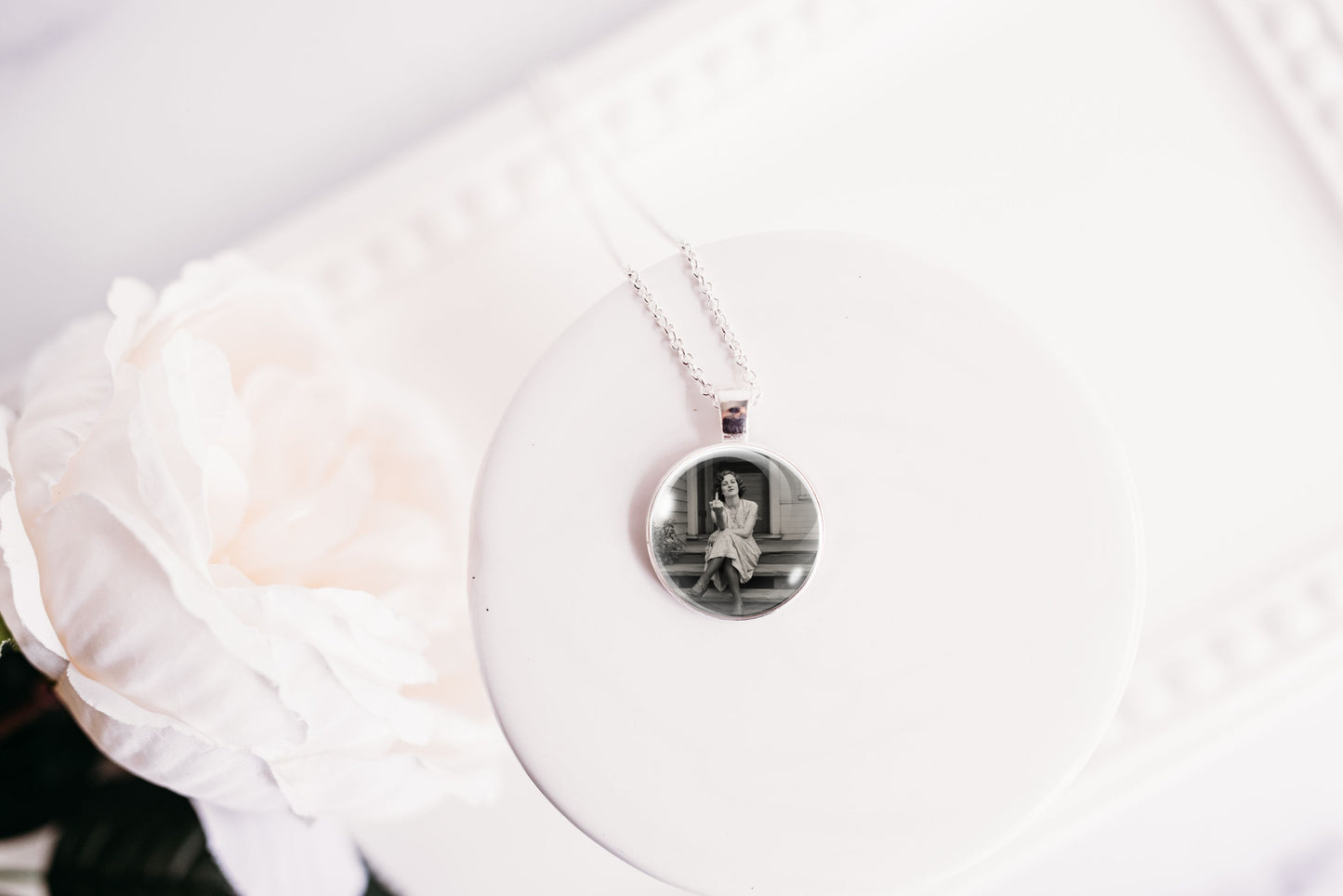 A silver necklace with a round photo pendant displayed on a white circular stand next to a white rose.