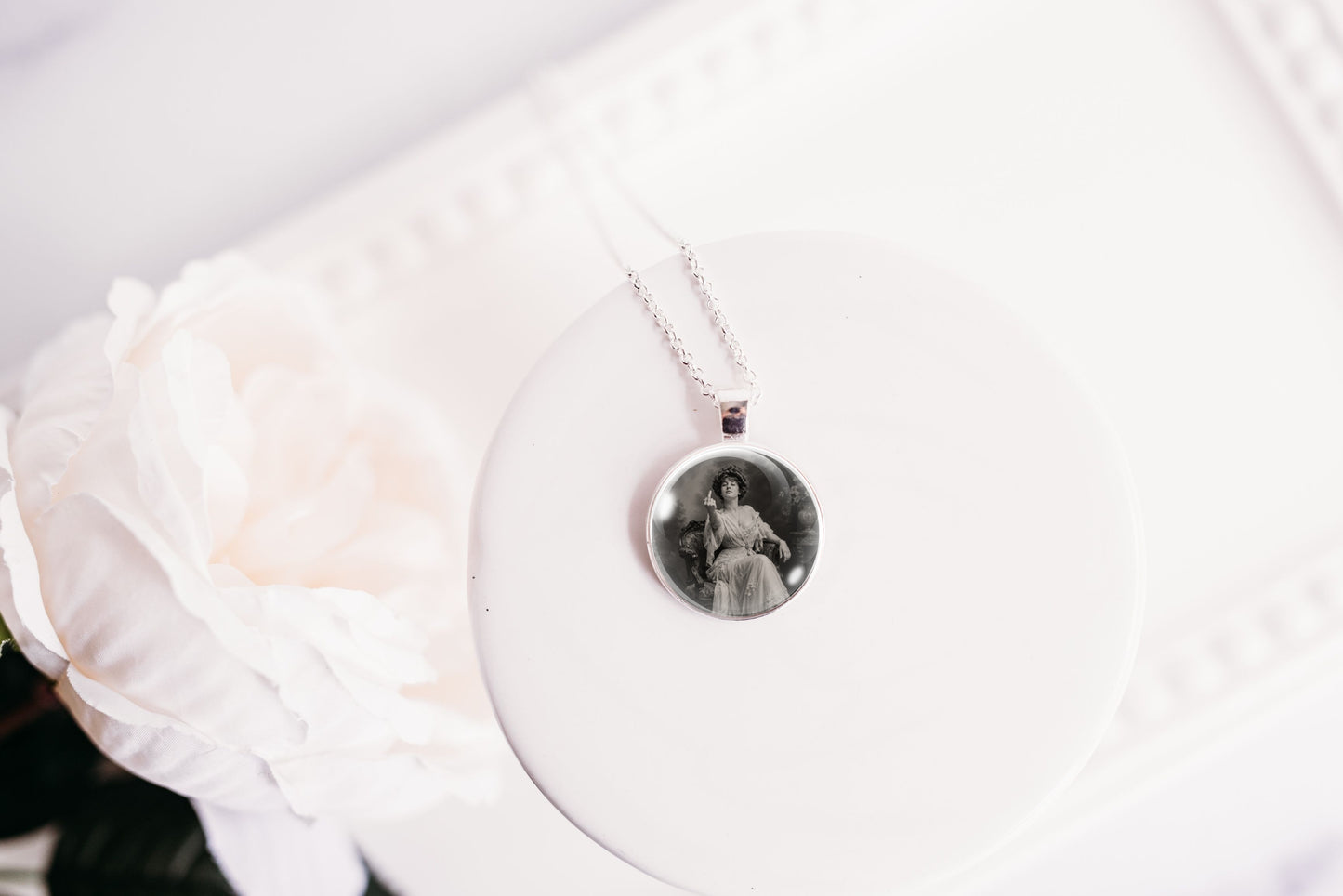 A silver-toned pendant necklace with a circular black-and-white portrait photo, displayed on a white round stand next to a white rose.