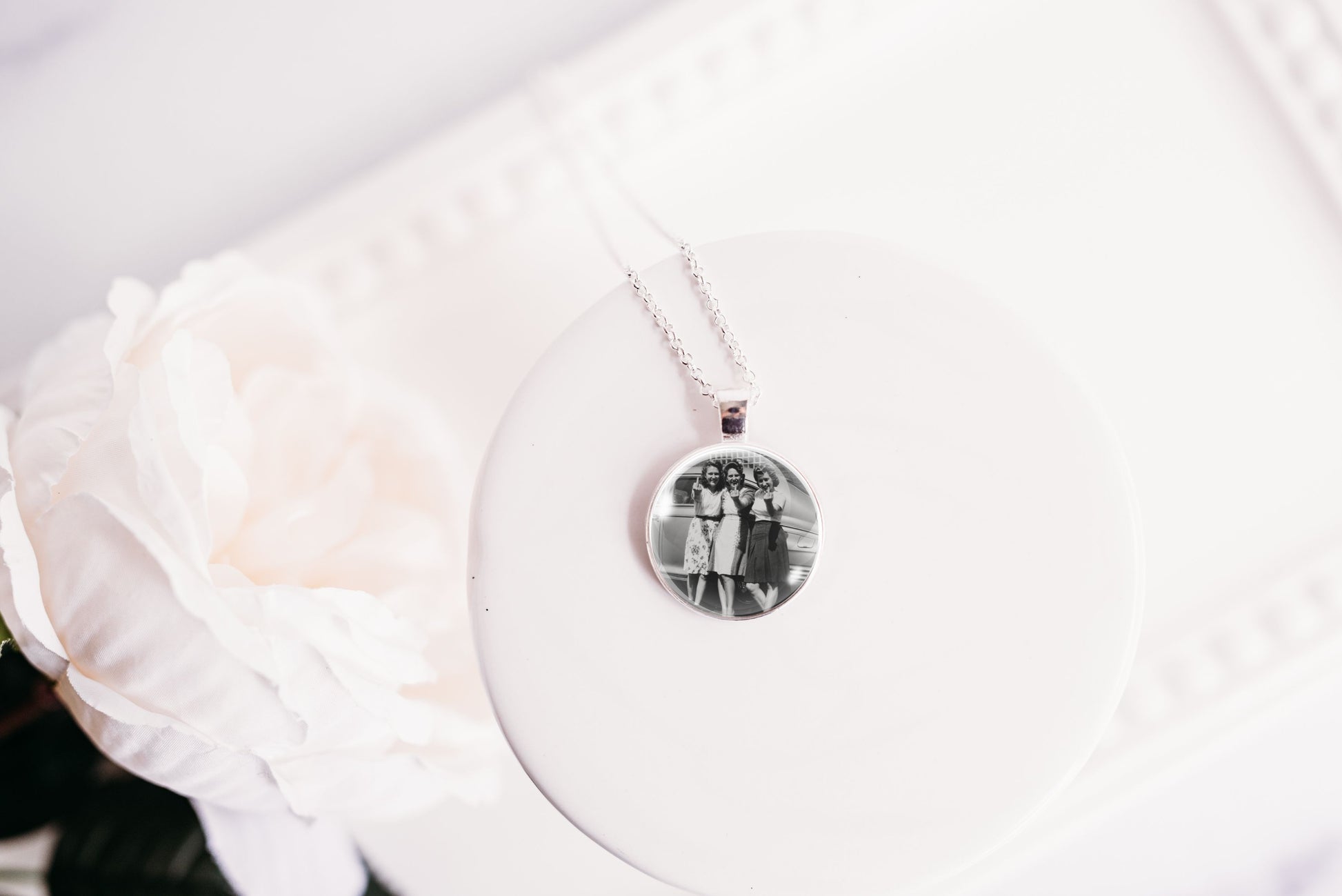 A round pendant necklace with a black-and-white photograph under a clear dome, hanging from a silver-toned chain on a white display stand with a soft white flower in the background.