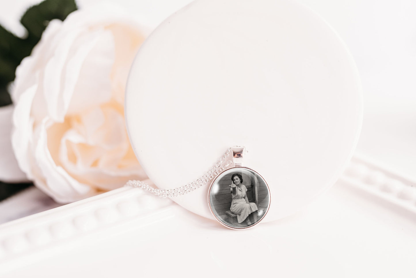 A silver-toned necklace with a round pendant displaying a black-and-white portrait photo, resting on a white display stand next to a soft white rose.