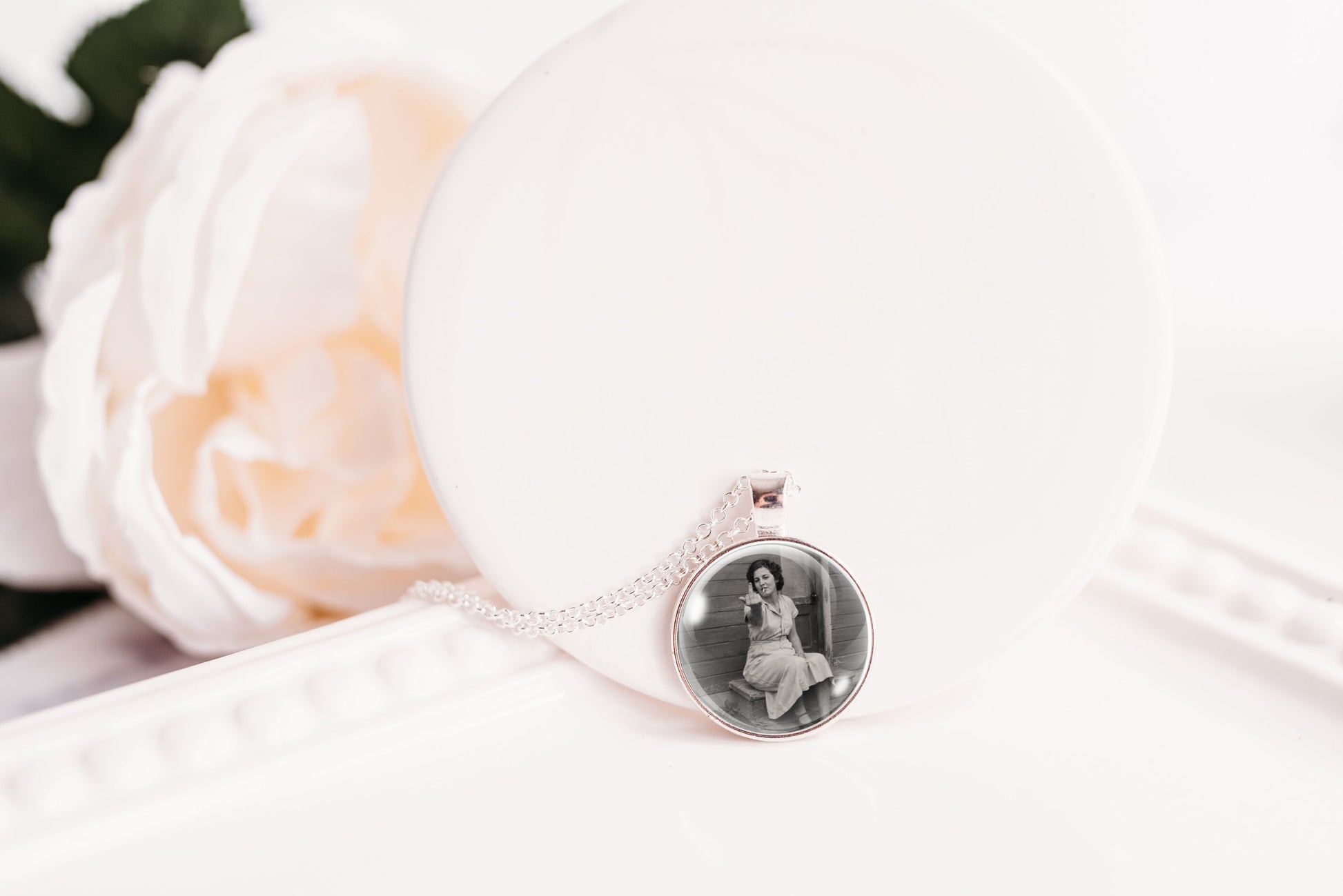 A silver-toned necklace with a round pendant displaying a black-and-white portrait photo, resting on a white display stand next to a soft white rose.