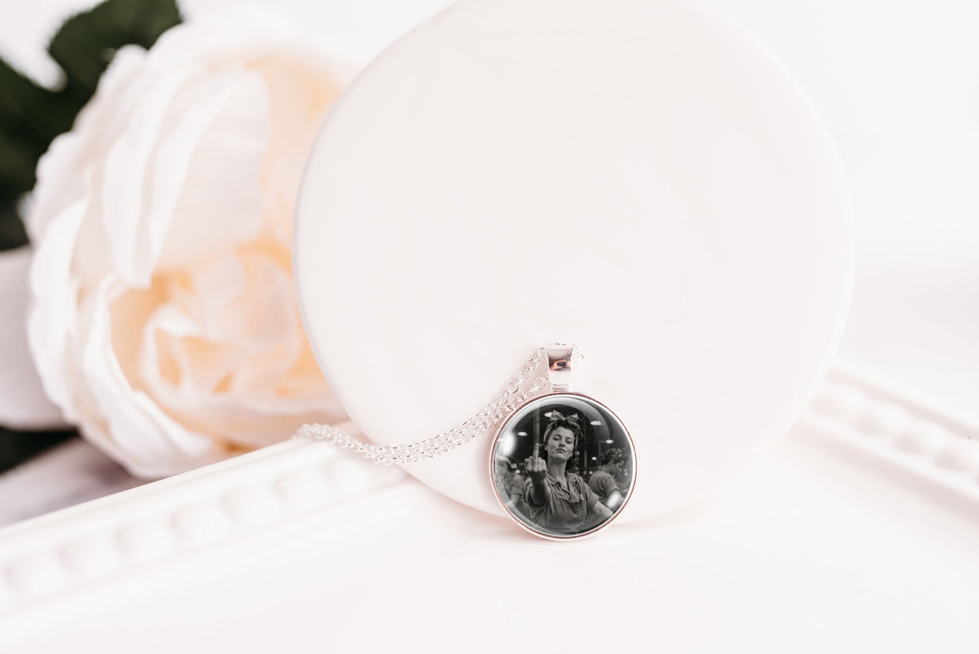 A silver-toned necklace with a round pendant displaying a black-and-white portrait photo, resting on a white display stand next to a soft white rose.