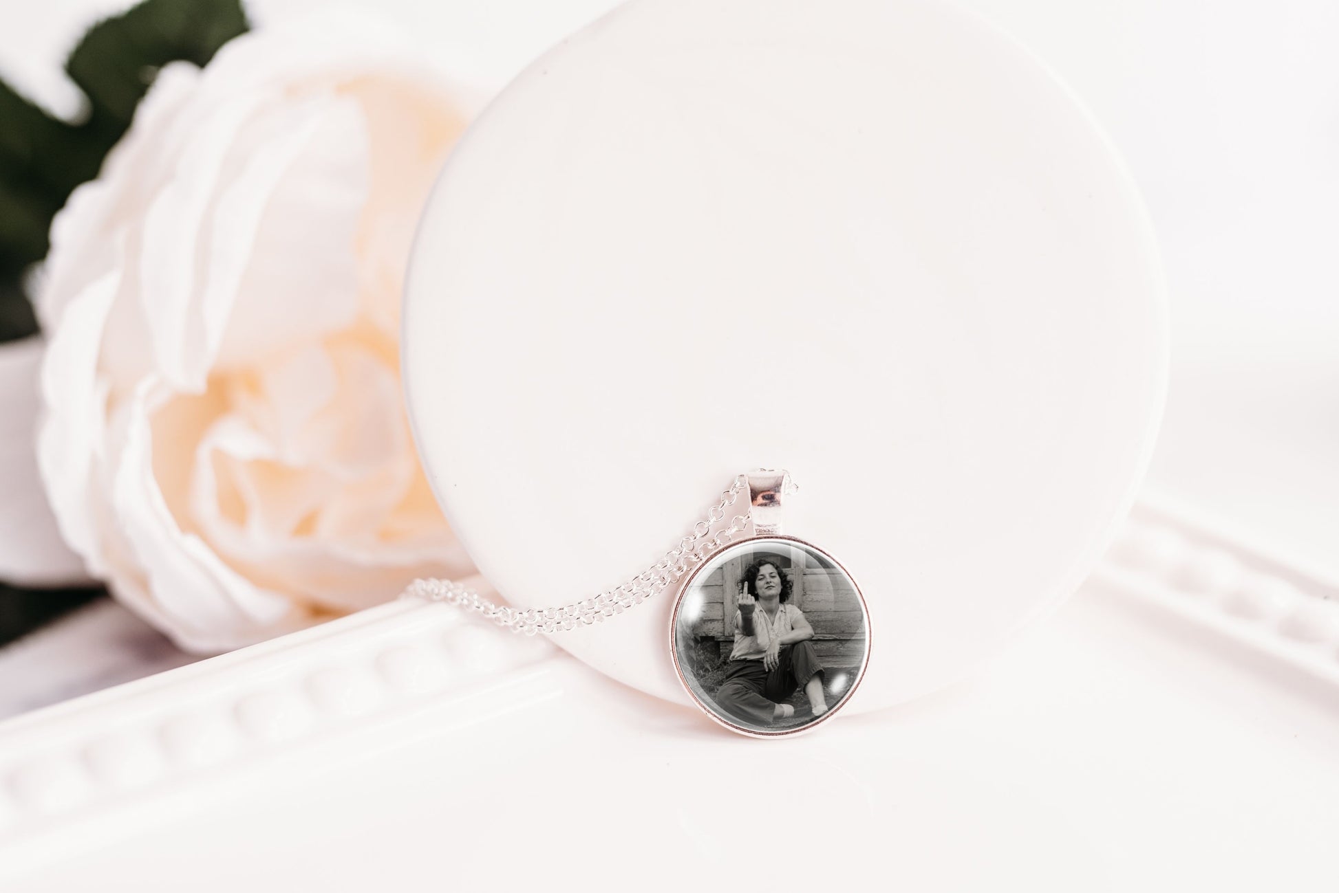 A silver-toned necklace with a round pendant displaying a black-and-white photo of a woman, resting on a white display stand next to a soft white rose.