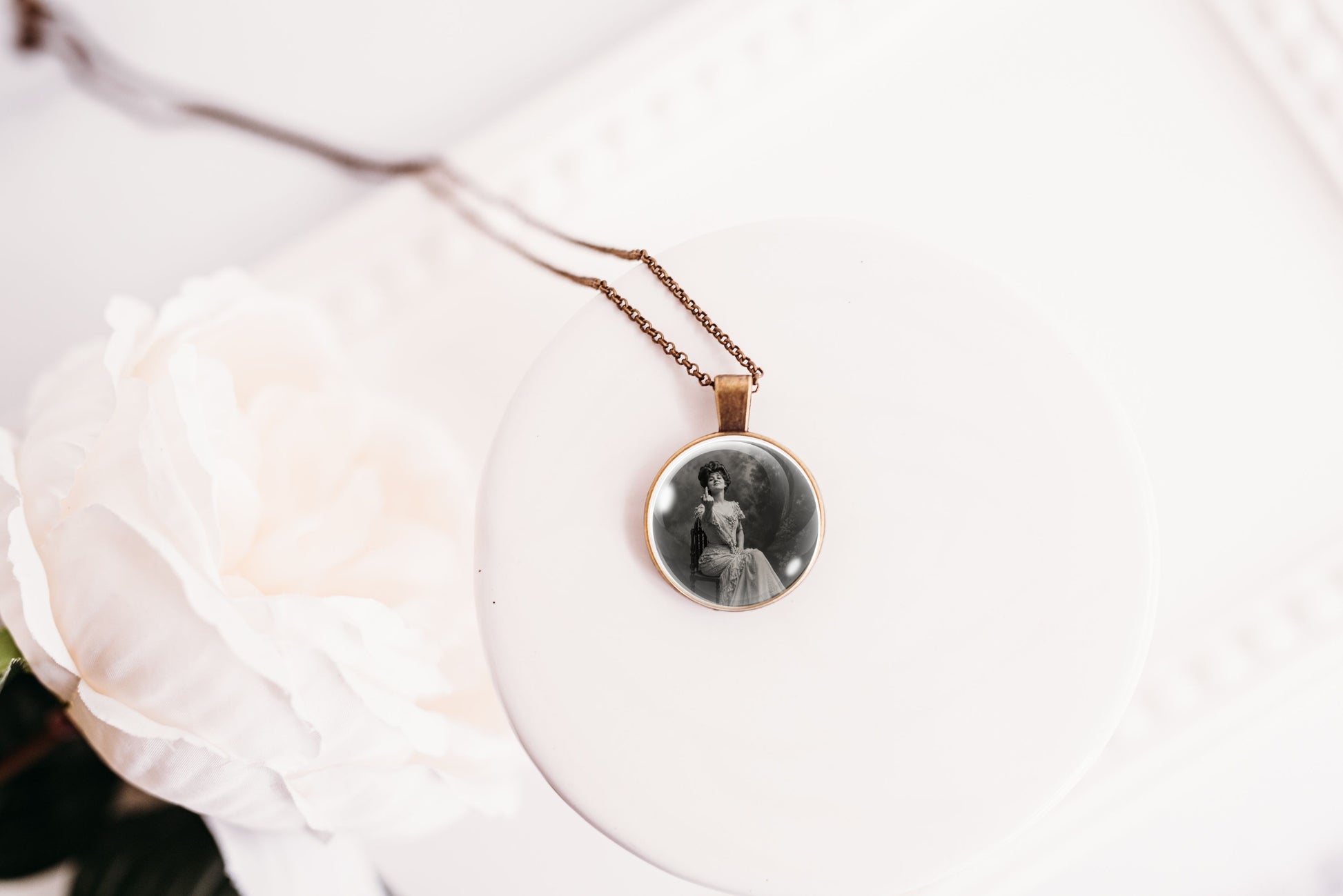 A round pendant necklace with a black-and-white portrait photo in a bronze-toned bezel, hanging from a delicate chain on a white display stand beside a white rose.