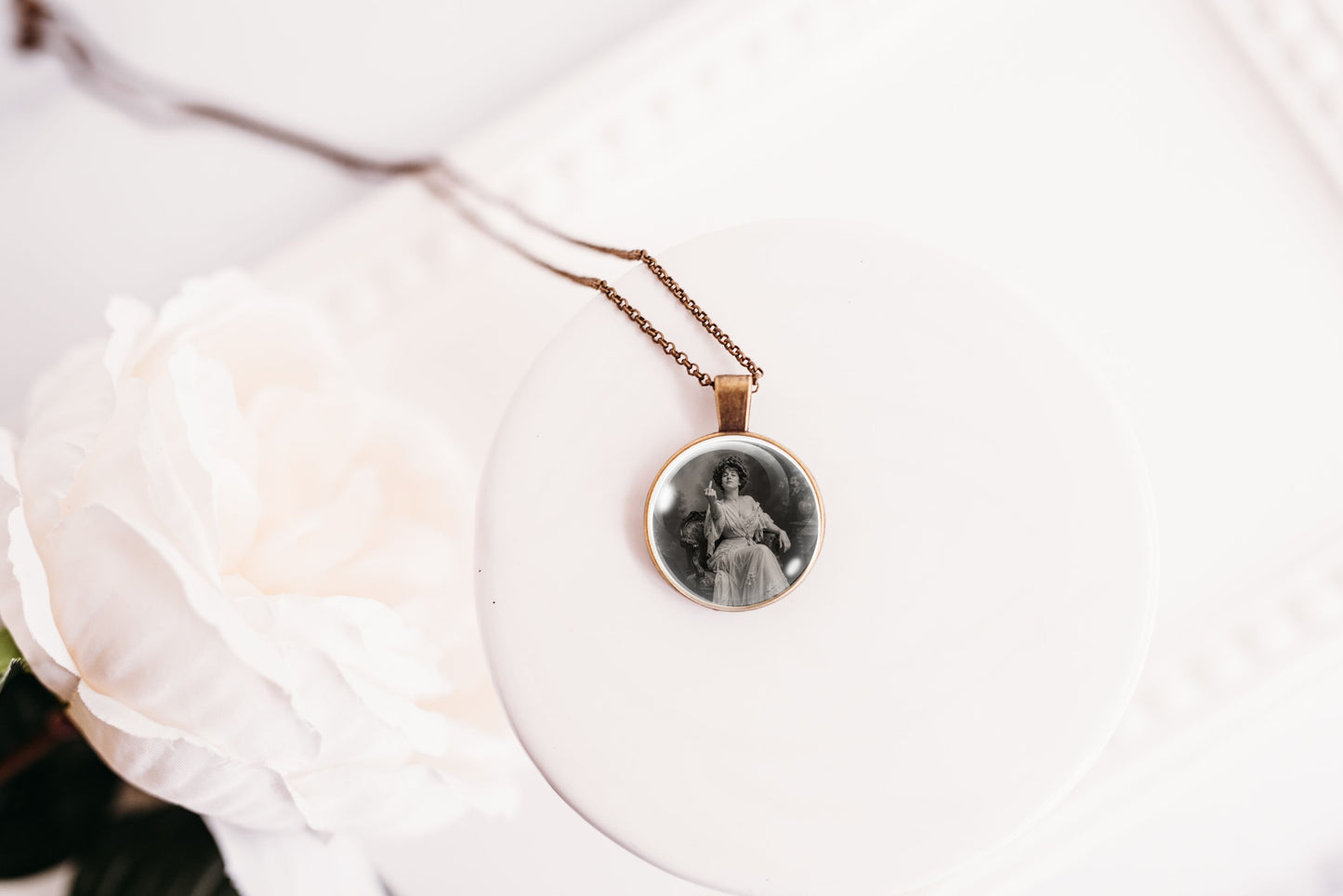 A round pendant necklace with a vintage black-and-white portrait photo, hanging from a delicate chain on a white display stand next to a white rose.