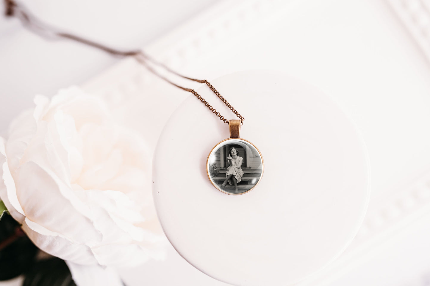 A close-up of a necklace with a round pendant showing a black-and-white portrait photo, resting on a white circular display with a soft white flower in the foreground.