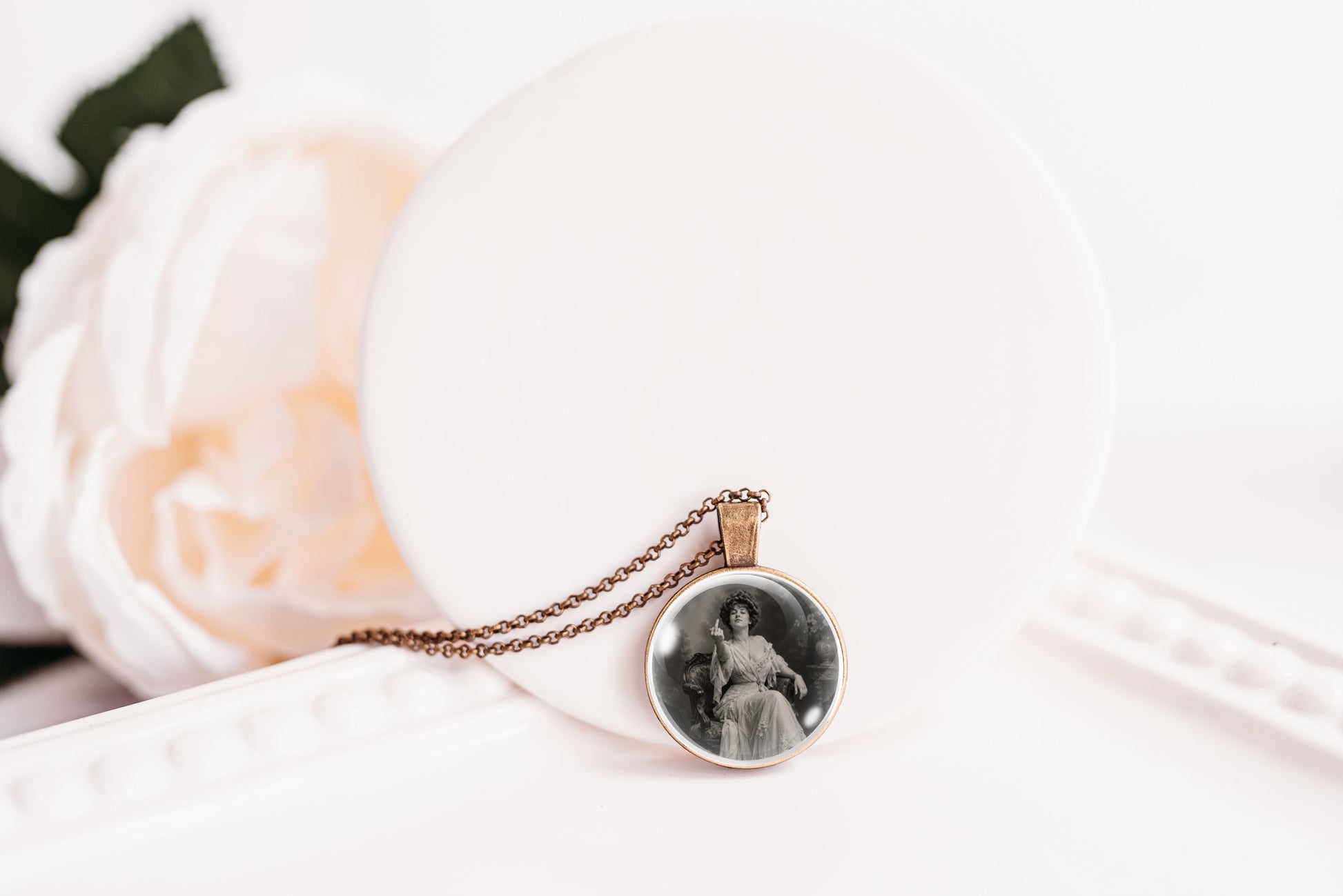 A round pendant necklace with a vintage black-and-white portrait photo, hanging from a bronze-toned chain and displayed on a white circular stand with a soft white flower in the background.