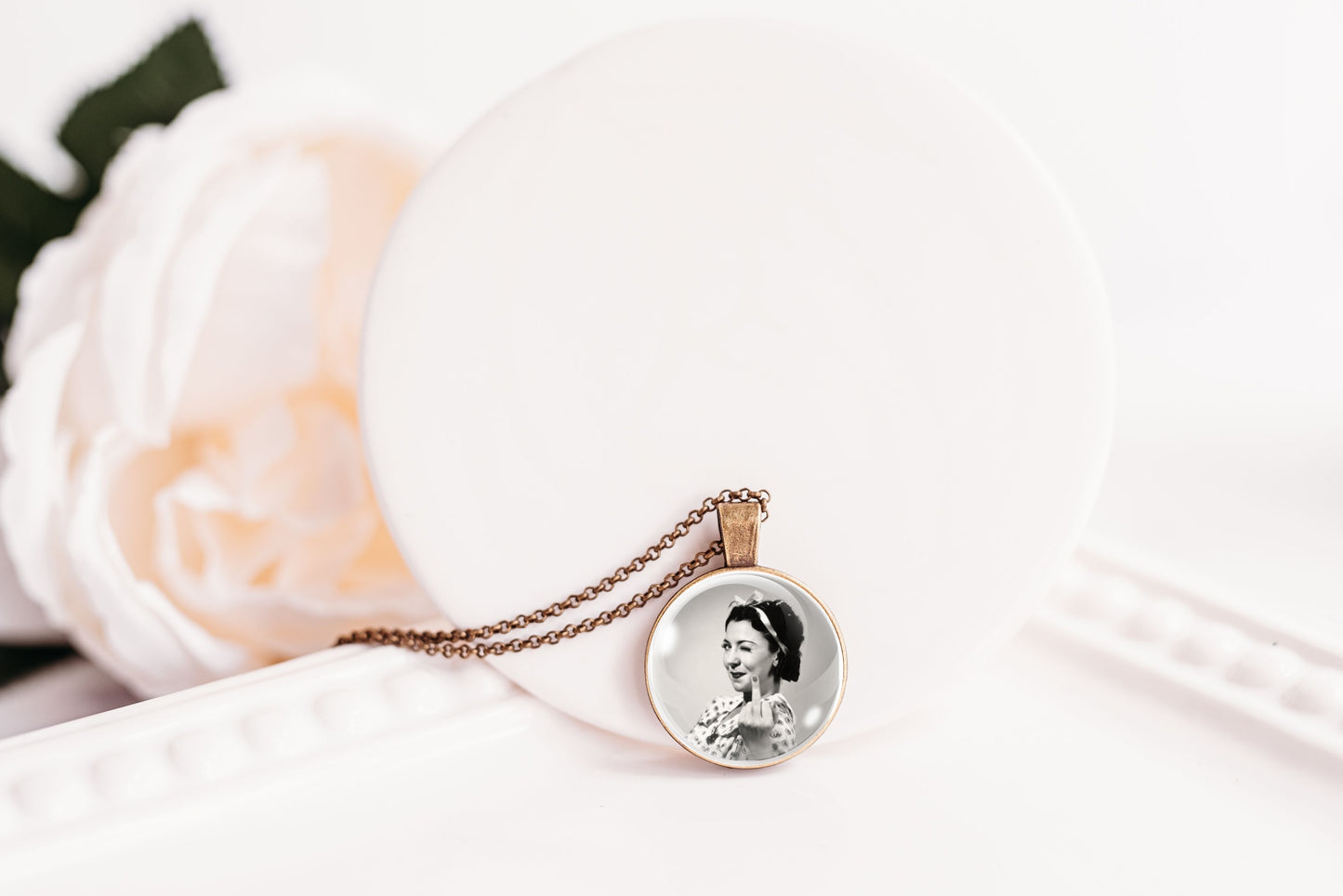 A round pendant necklace featuring a black and white portrait photo of a woman, suspended from a bronze-toned chain, displayed on a white circular stand next to a soft white rose.