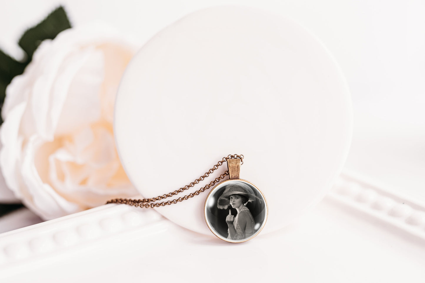 A round pendant necklace with a black-and-white printed photo in a bronze-plated bezel, hanging from a bronze chain on a white display stand with a soft white flower prop.