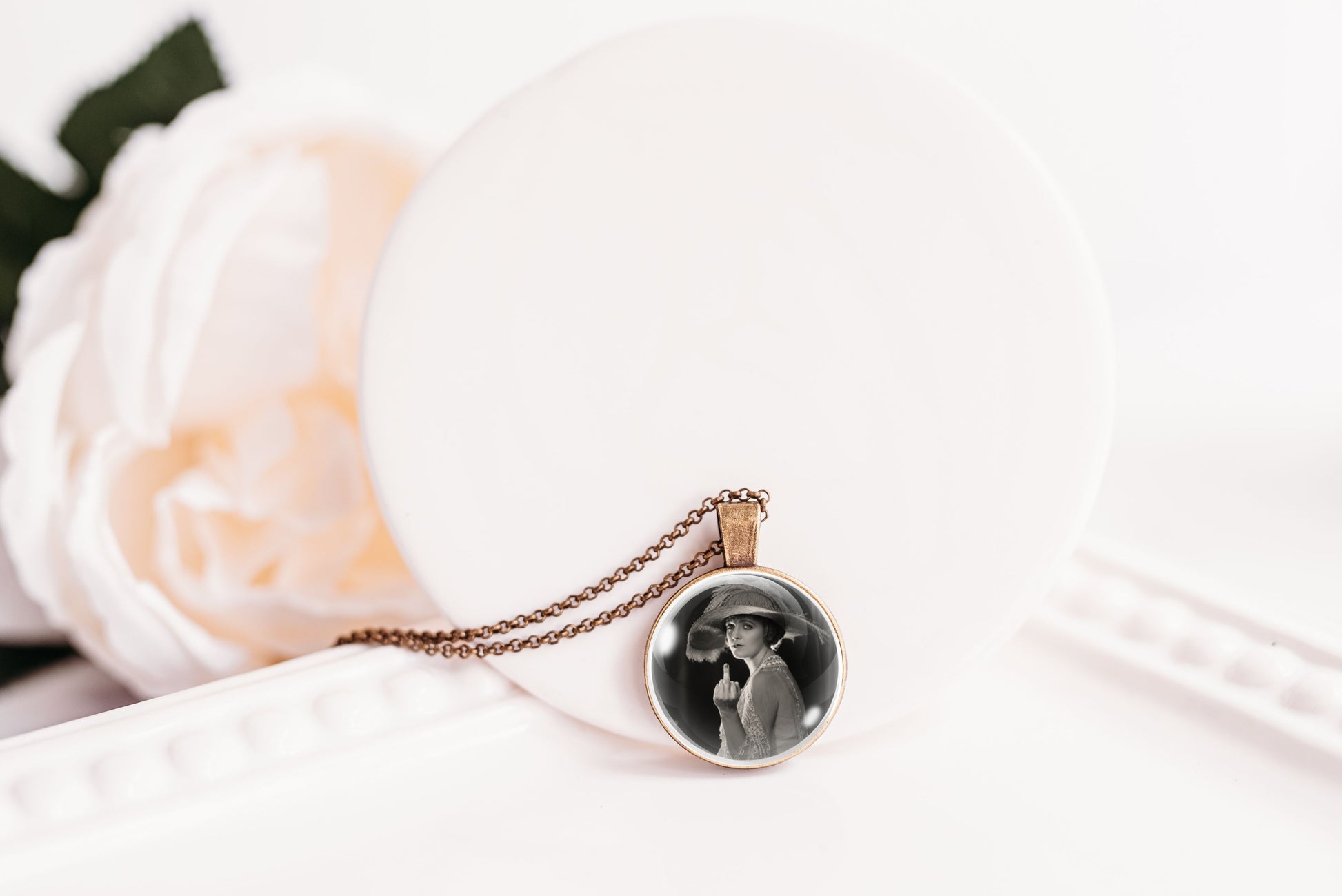 A round pendant necklace with a black-and-white printed photo in a bronze-plated bezel, hanging from a bronze chain on a white display stand with a soft white flower prop.