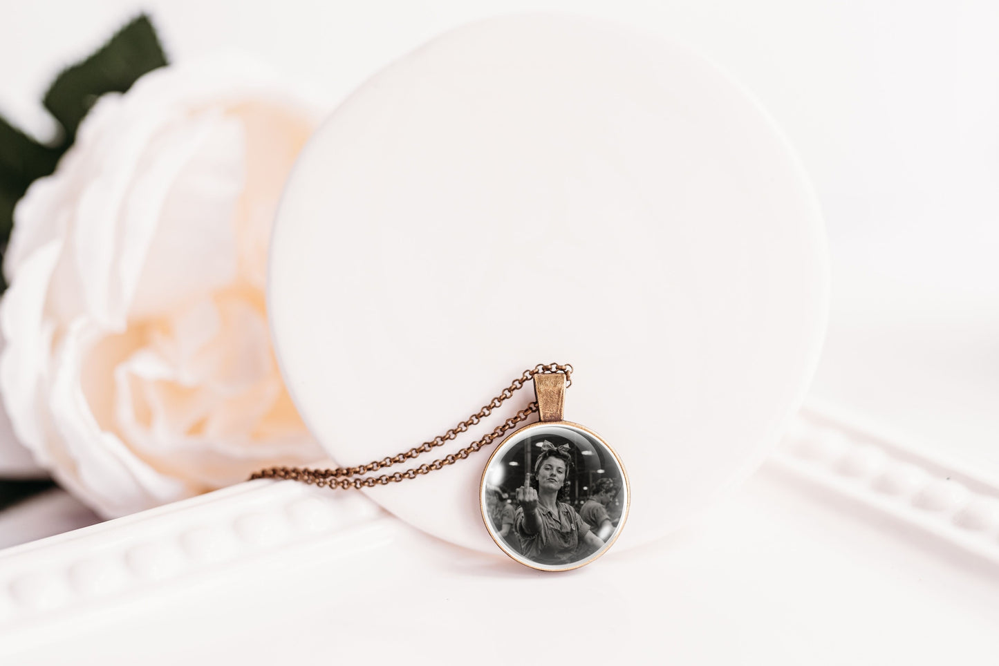 A round pendant necklace with a black-and-white printed photo in a silver-toned bezel, hanging from a bronze-toned chain on a white display stand with a soft white flower prop.