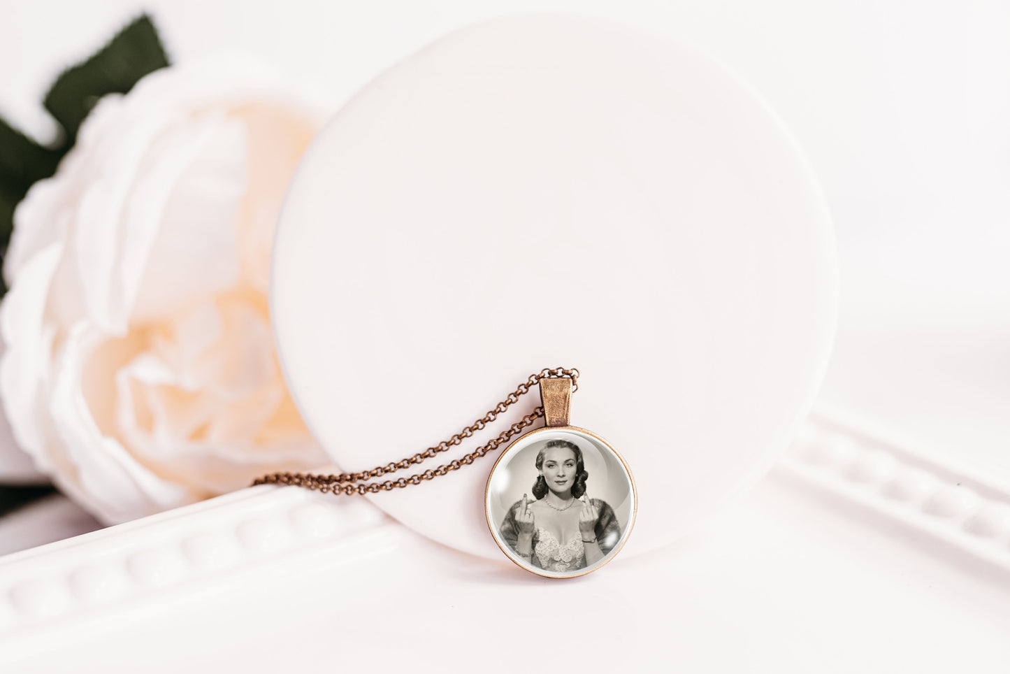 A round pendant necklace with a vintage-style black and white portrait photo, hanging from a delicate bronze-toned chain on a white display stand with a soft white flower in the background.